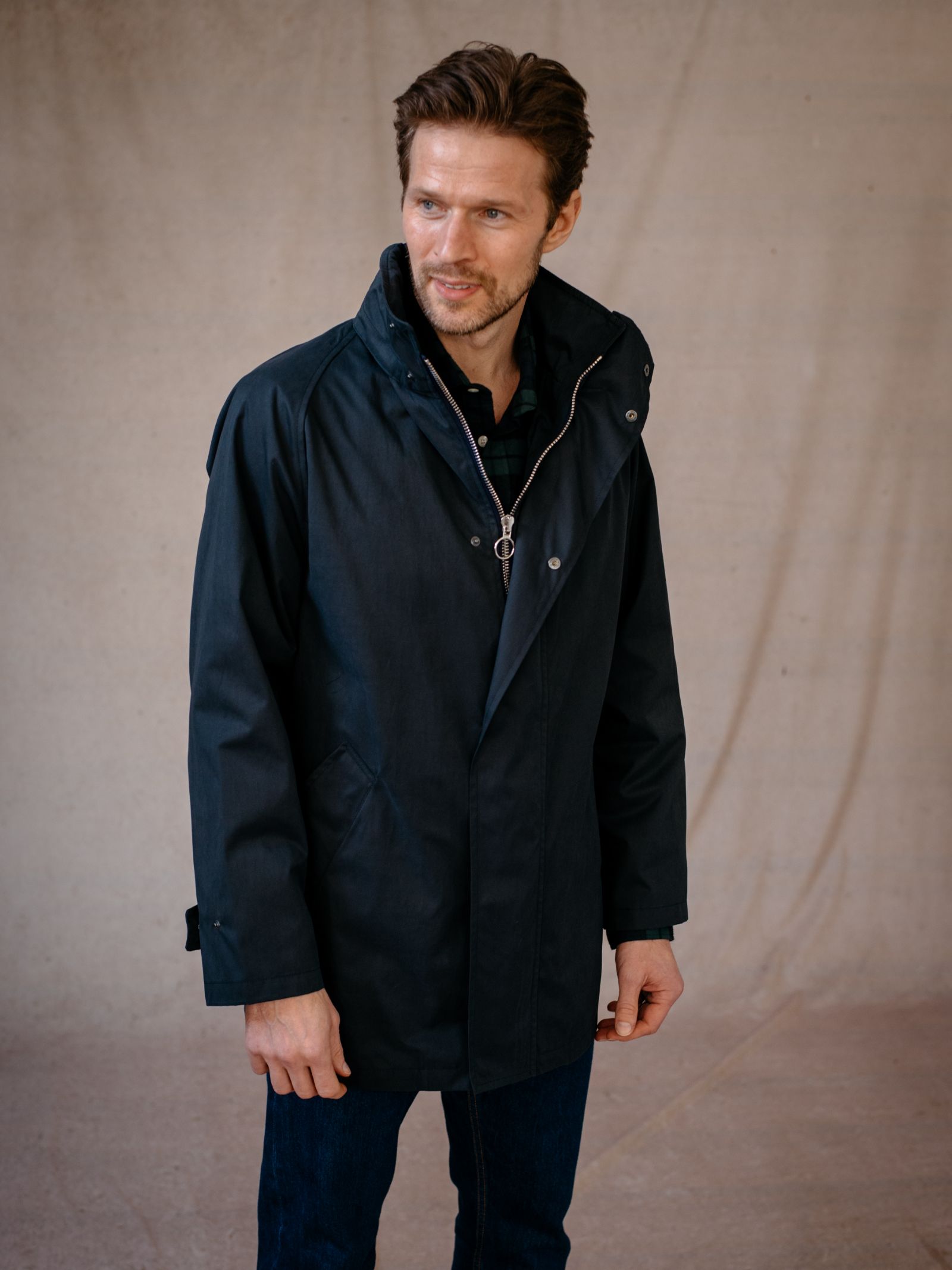 A man with light skin and brown hair wears a dark blue Campbells of Beauly Mont Coat over a dark shirt and blue jeans, standing against a beige background and looking slightly left with a subtle smile.