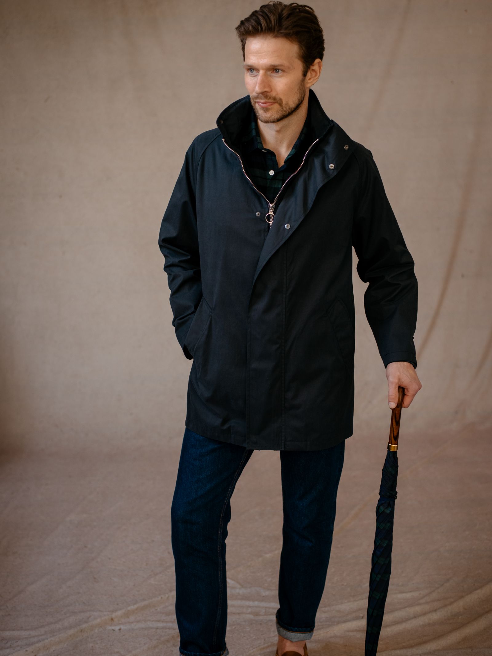A man with short brown hair and a trimmed beard wears the Campbells of Beauly Mont Coat over a black shirt and blue jeans, standing on a tan floor and holding a closed umbrella with a wooden handle, gazing slightly left with a neutral expression.