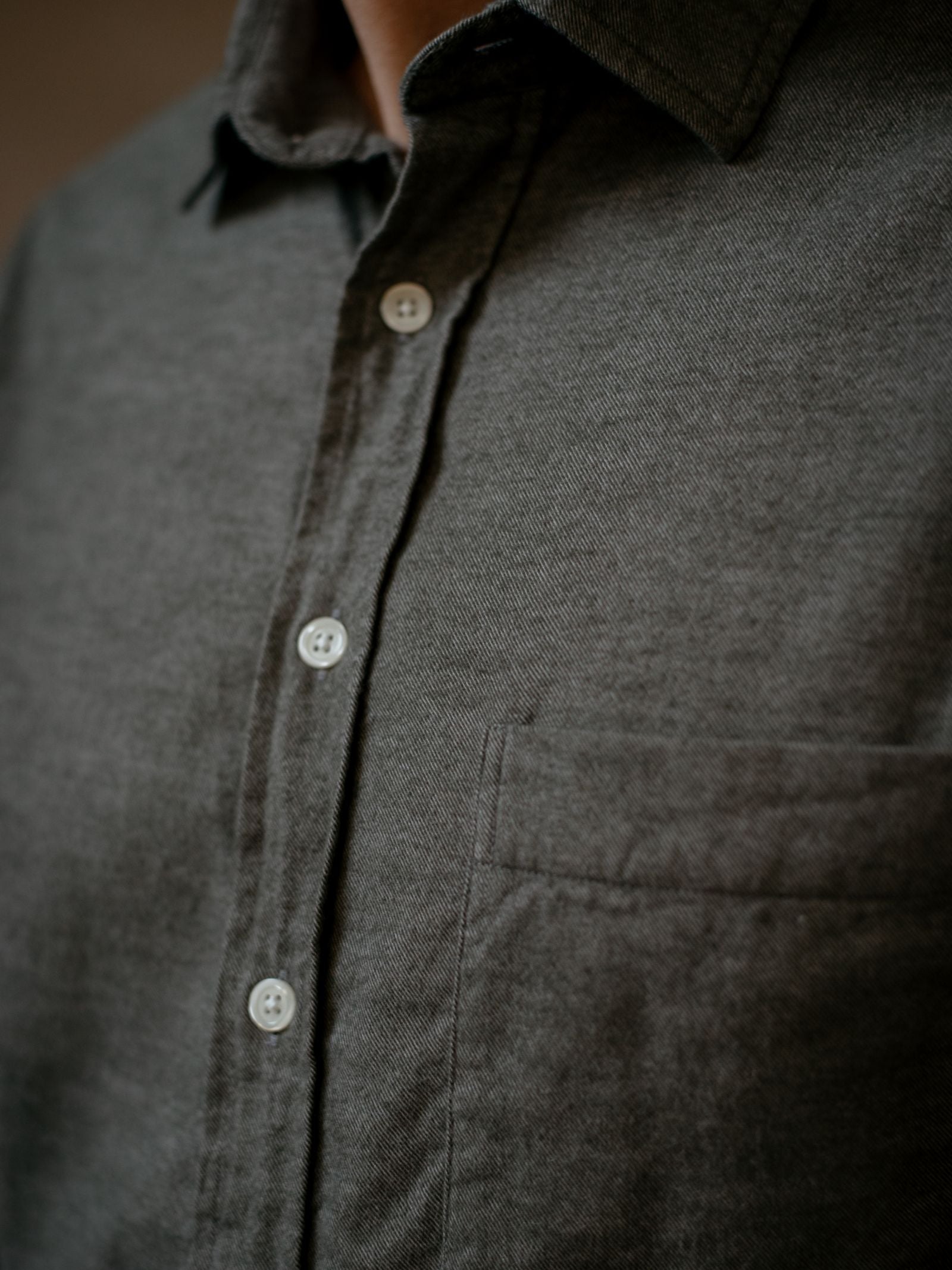 Close-up of a person wearing the Campbells of Beauly Twill Shirt, a dark gray classic fit button-up in brushed cotton with white buttons and a visible left chest pocket. The soft focus highlights the textured fabric and collar.