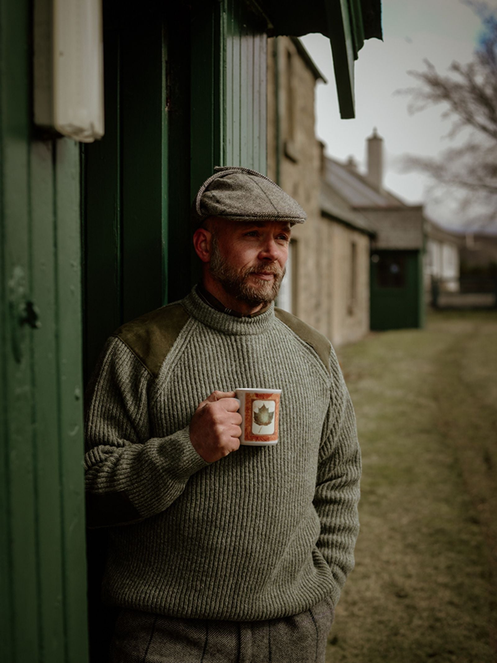 A man wearing Campbell’s of Beauly’s Scotia Suede Jumper and a flat cap leans against a green building, mug in hand, gazing thoughtfully into the distance amidst a rural backdrop of old stone buildings.