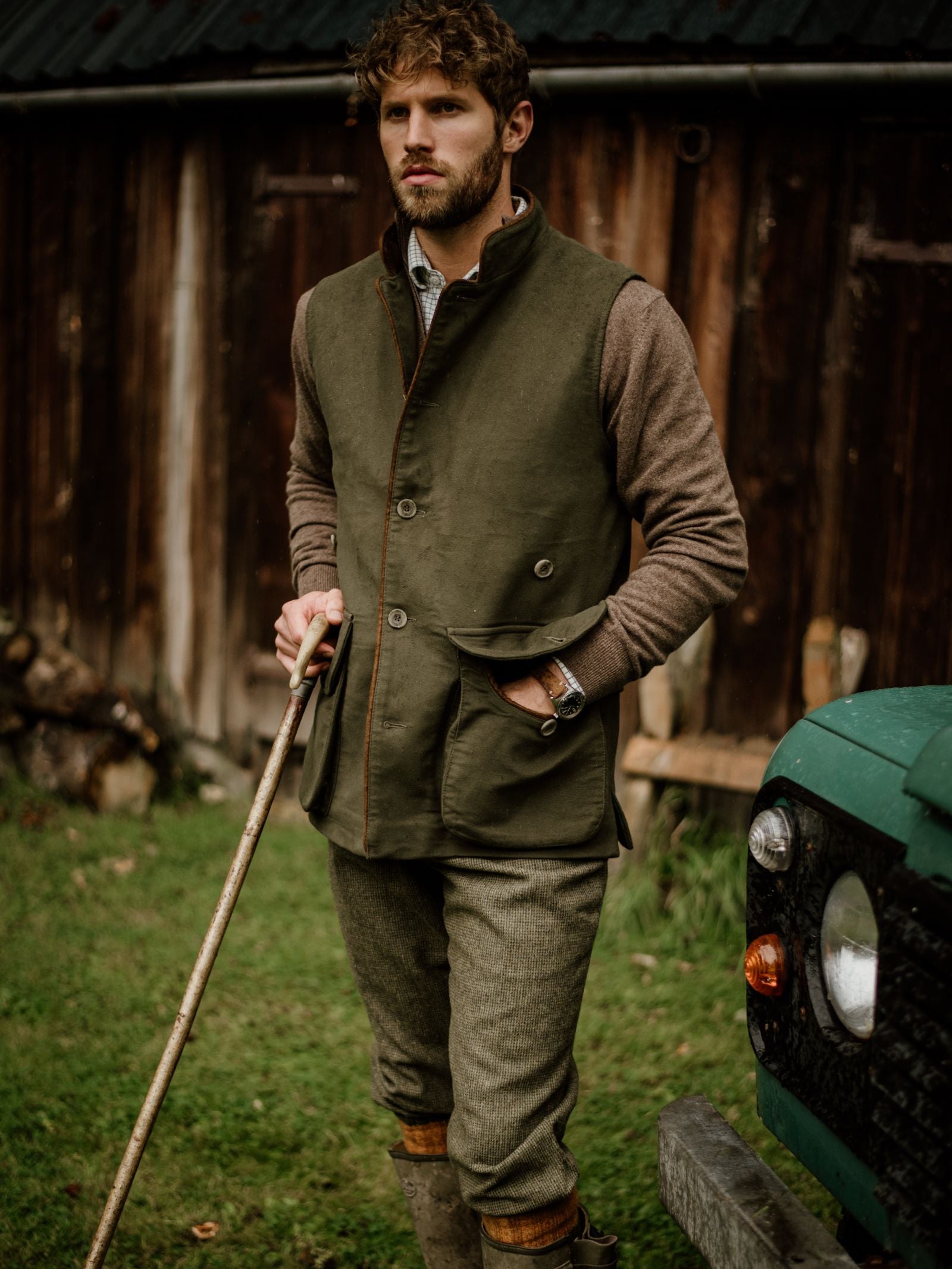 A man stands outdoors holding a wooden stick, wearing the Campbells of Beauly Field Nehru Gilet over a brown sweater and tweed pants with knee-high boots. A green vehicle and rustic wooden building appear in the background.