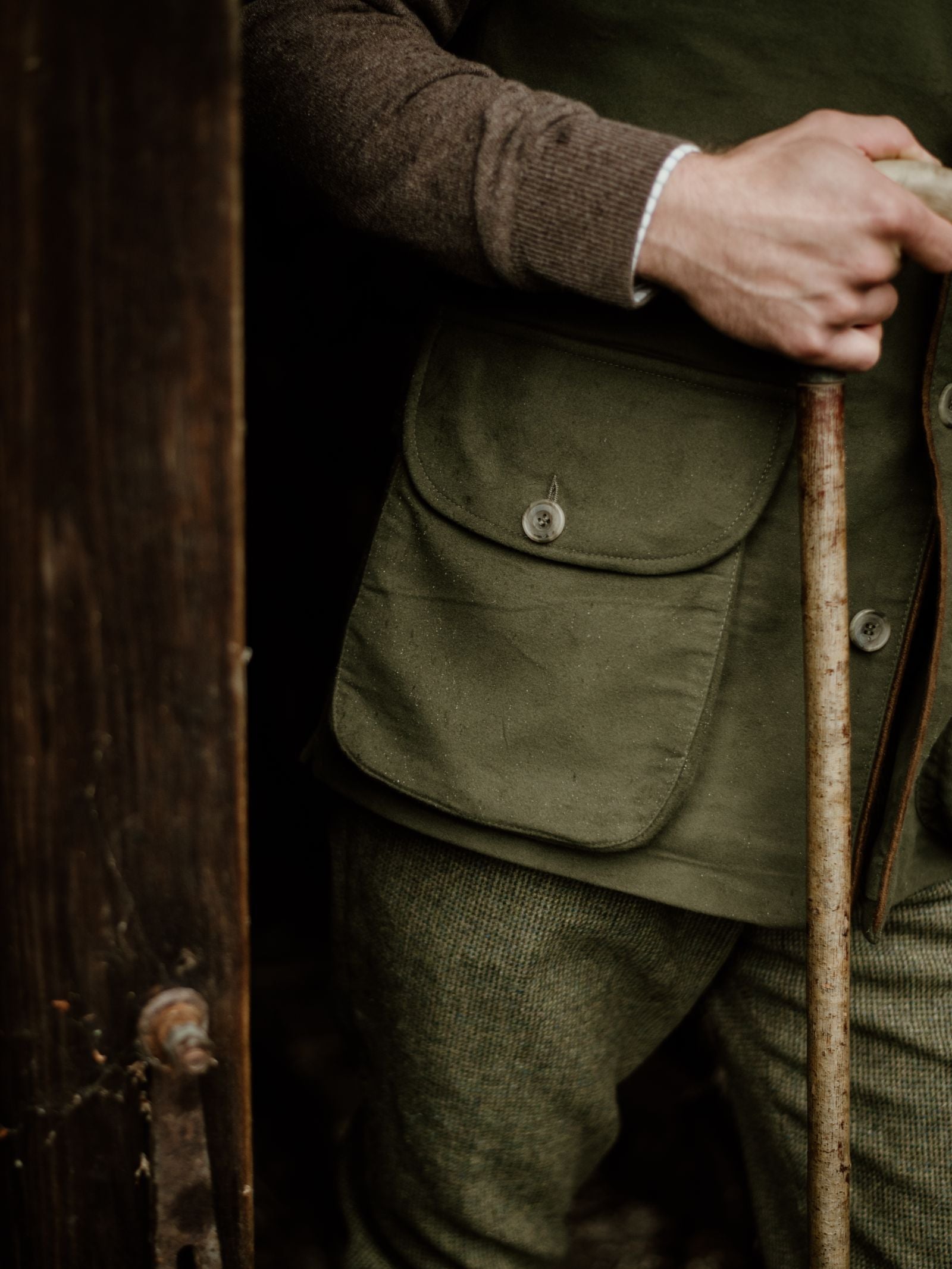 Wearing a Campbells of Beauly Field Nehru Gilet in olive green moleskin with quilted lining over a brown sweater, a person holds a wooden walking stick by a weathered door, evoking an outdoor or rustic atmosphere.