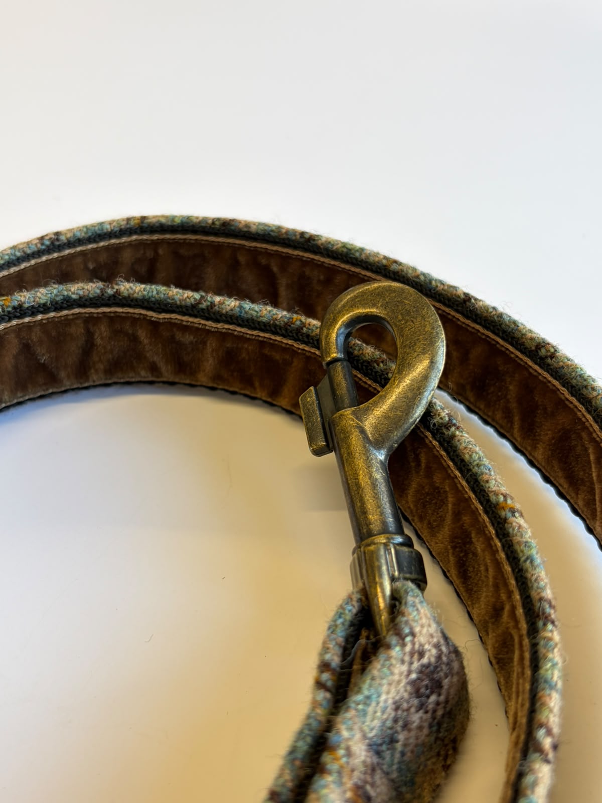 A close-up of the Campbell's of Beauly House Tweed Dog Lead, featuring brown and green fabric and a brass clip, coiled on a white surface.