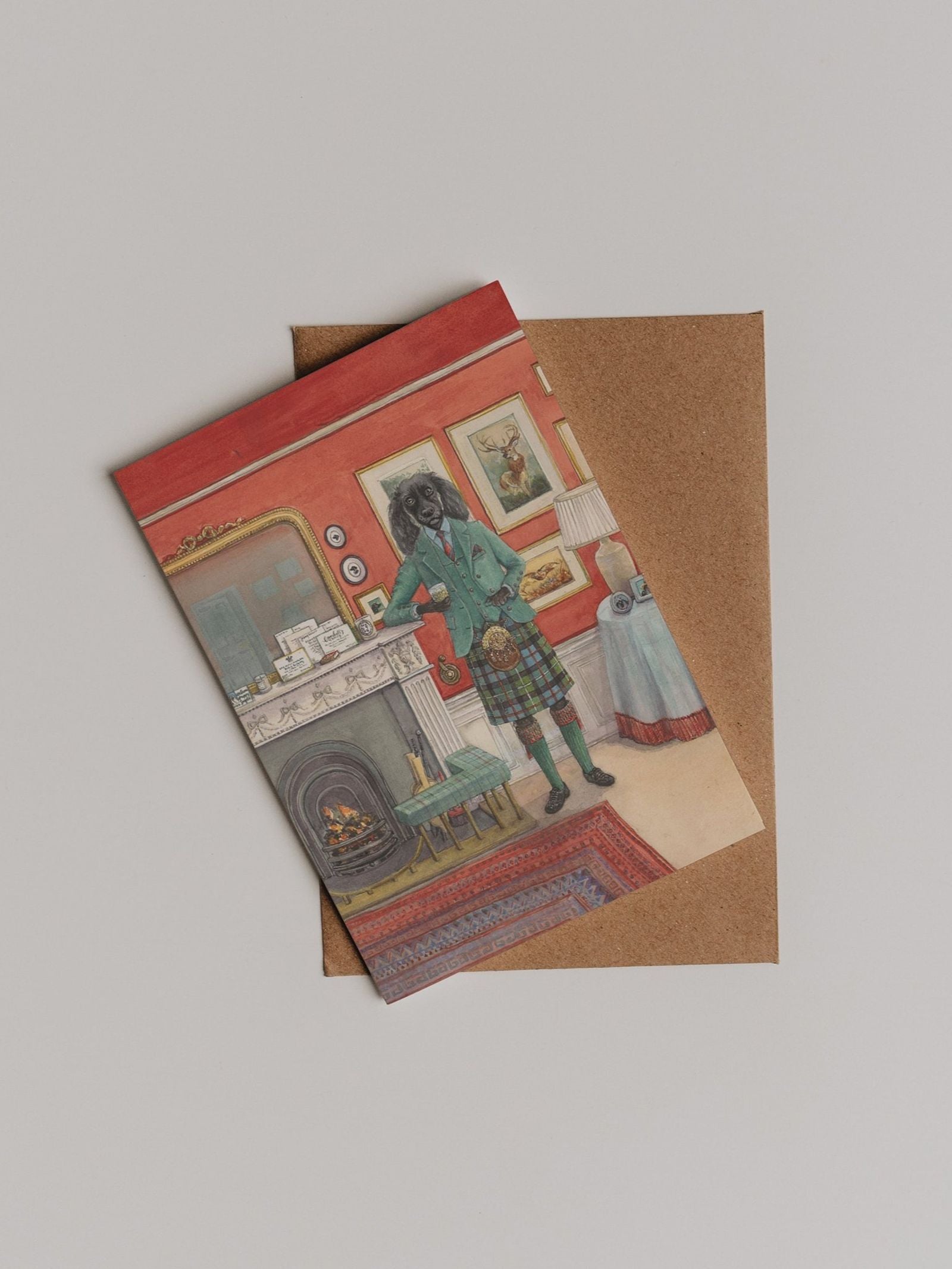 The Fireside Dram Greeting Card by Campbell’s of Beauly features Sophie Murray’s unique illustration of an anthropomorphic dog in a suit and kilt, standing in a vintage room with a fireplace and red walls. Presented on a brown envelope, this card makes the perfect thank you note.