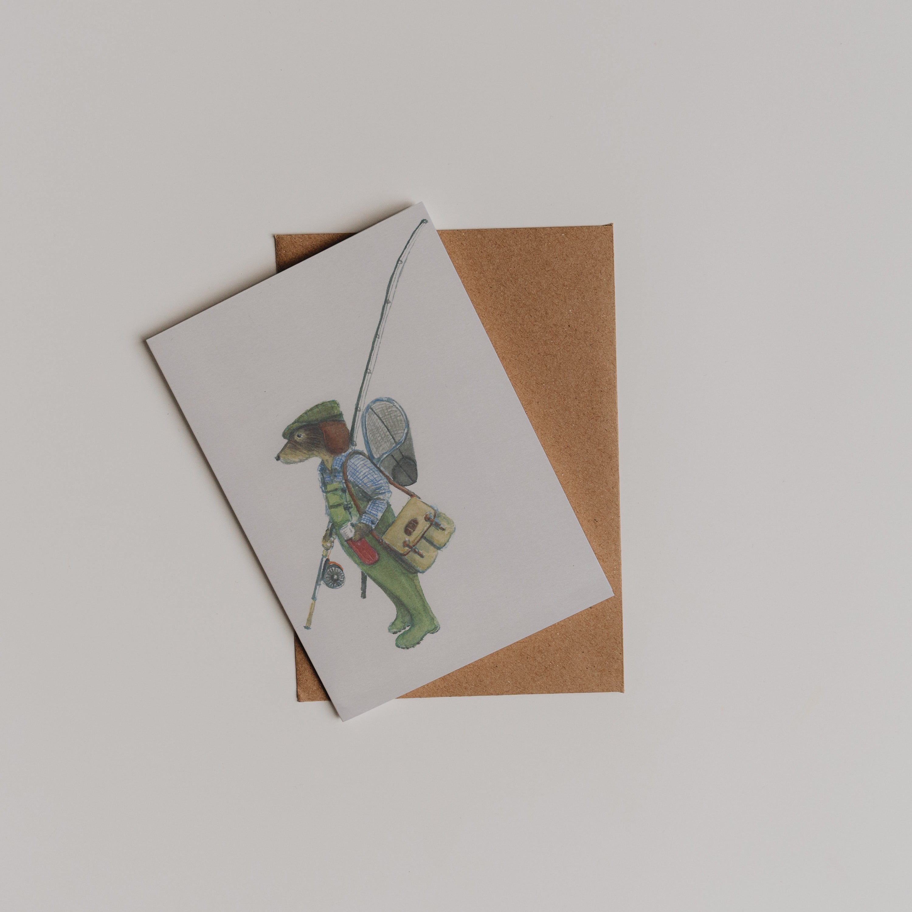 The Fishing Dachshund Greeting Card by Campbells of Beauly features a dog dressed as a fisherman with rod, net, and bags, partially atop a brown envelope on a white surface. Illustration by Sophie Murray.