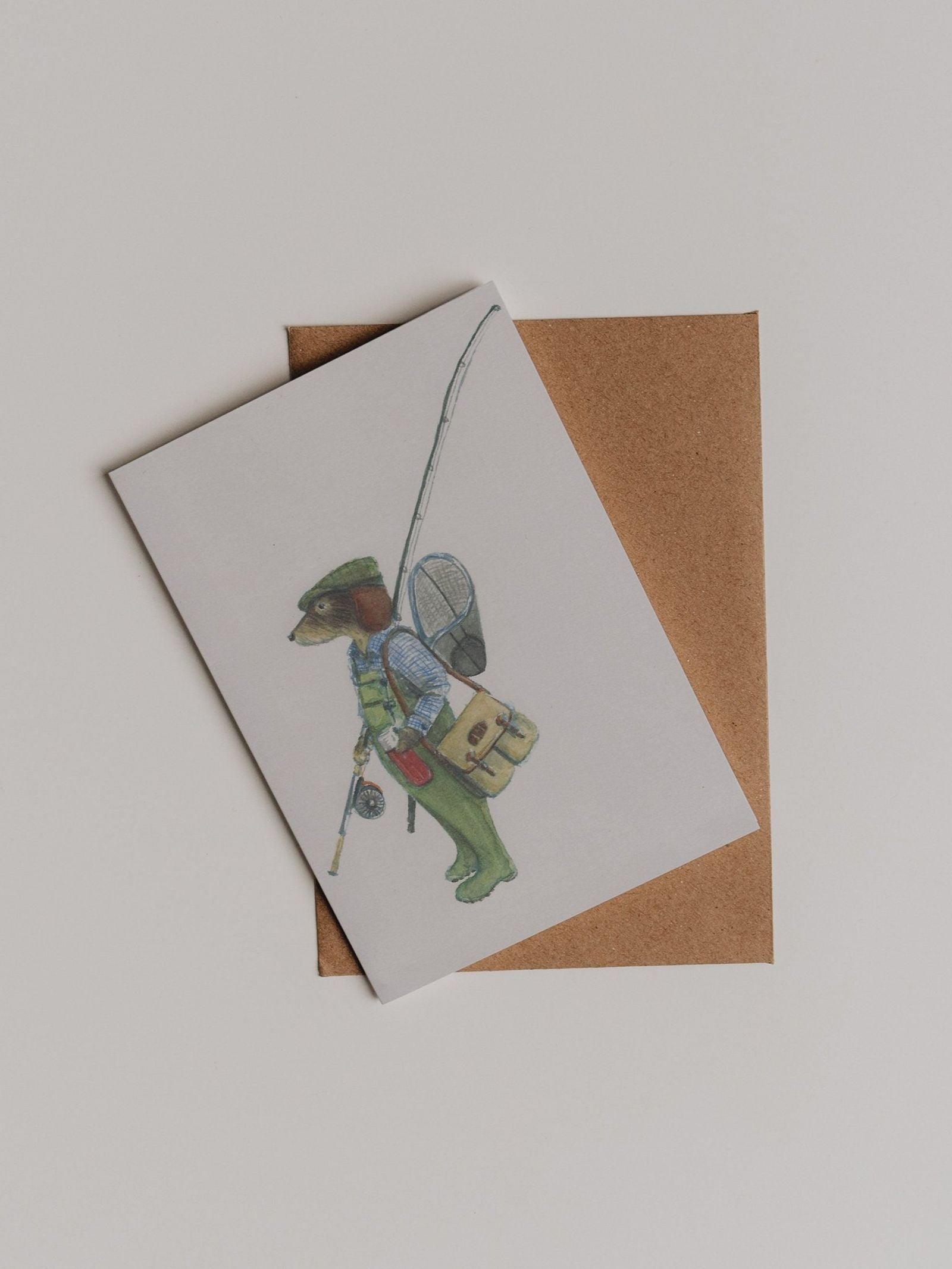 A charming “Fishing Dachshund Greeting Card” from Campbell’s of Beauly, featuring an illustration by Sophie Murray of an anthropomorphic dog dressed as a fisherman with a fishing rod, leaning on a brown envelope against a white background.
