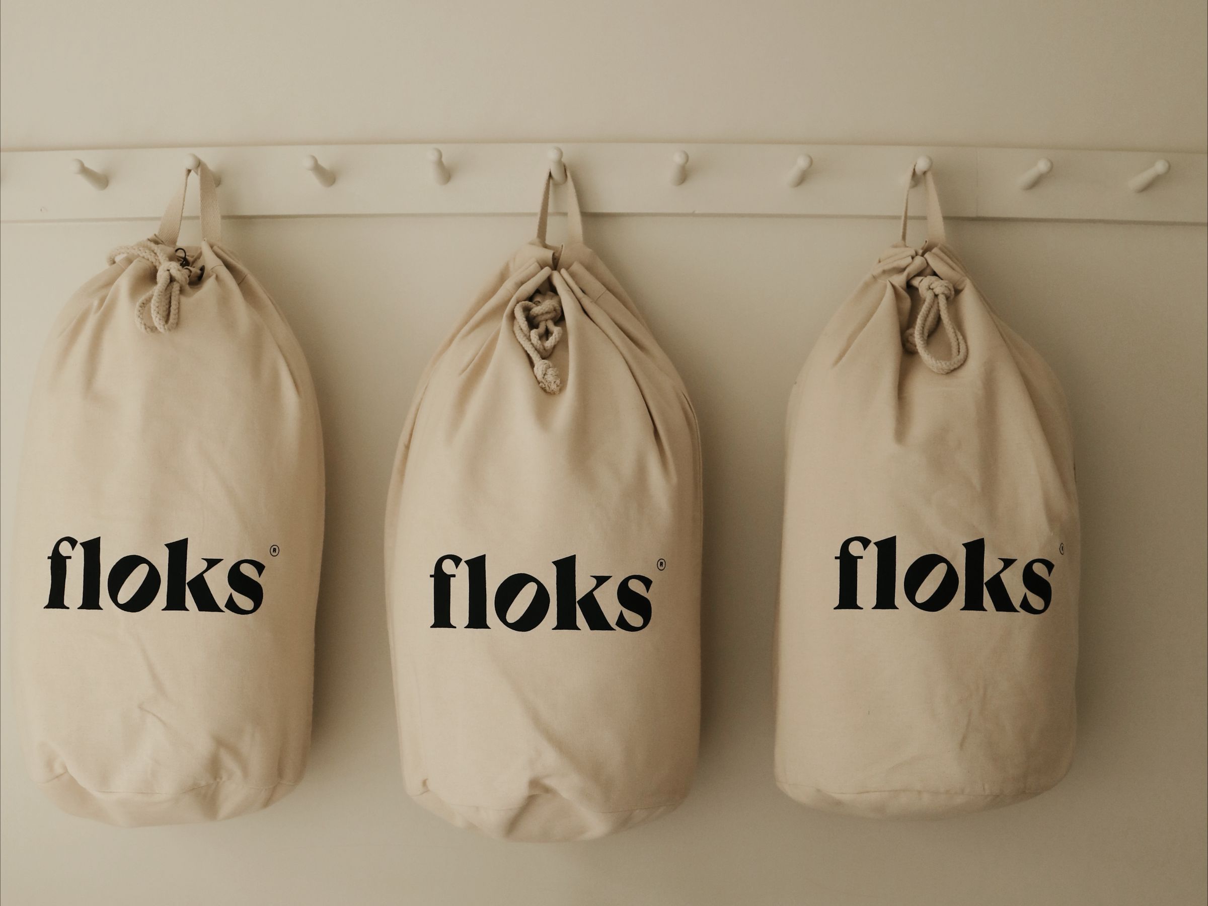Three beige drawstring bags labeled floks hang on white hooks against a light-colored wall. The bags are evenly spaced and identical in appearance.