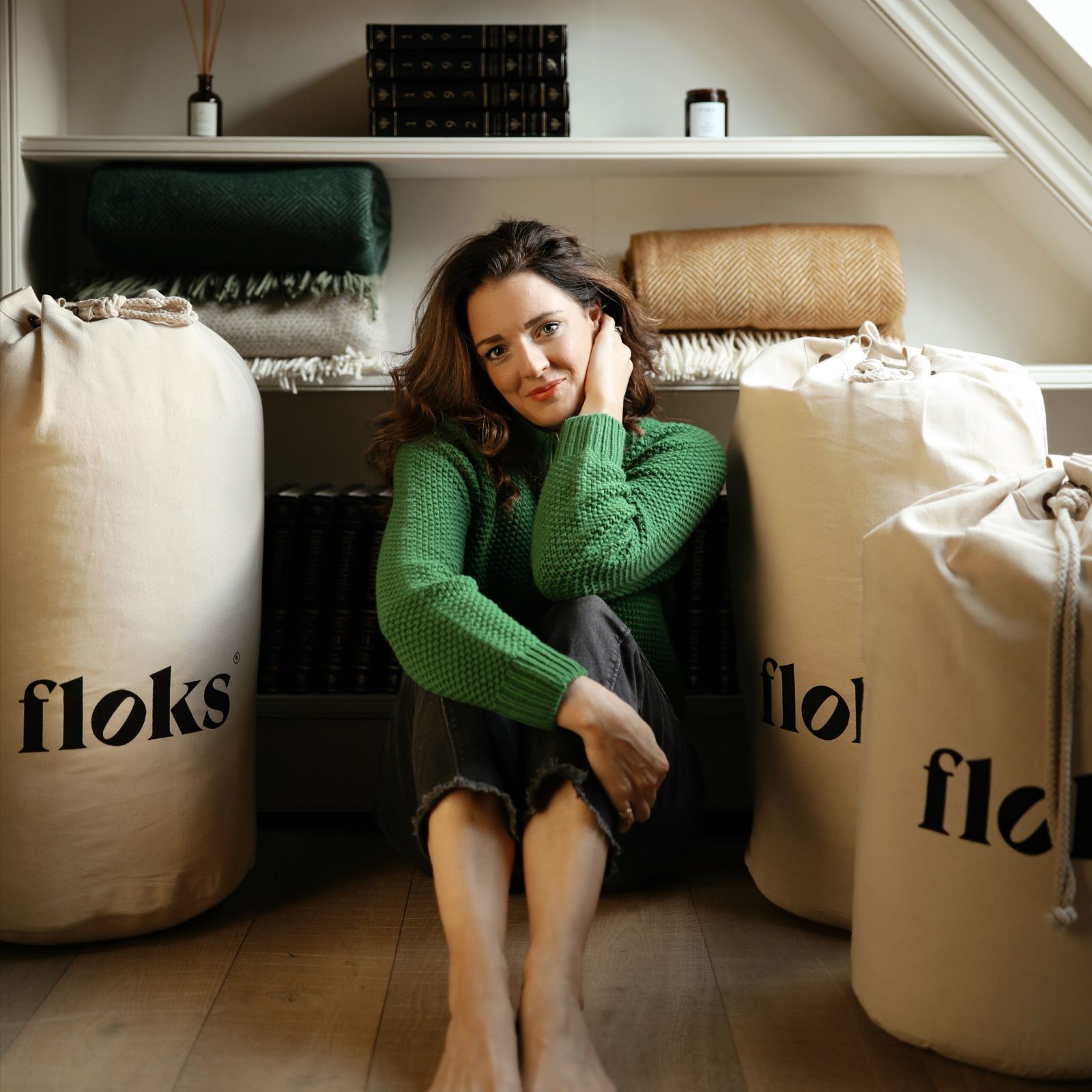 A woman in a green sweater sits barefoot on a wooden floor, leaning against a shelf with folded blankets and books. Large canvas bags labeled floks are placed on either side of her.