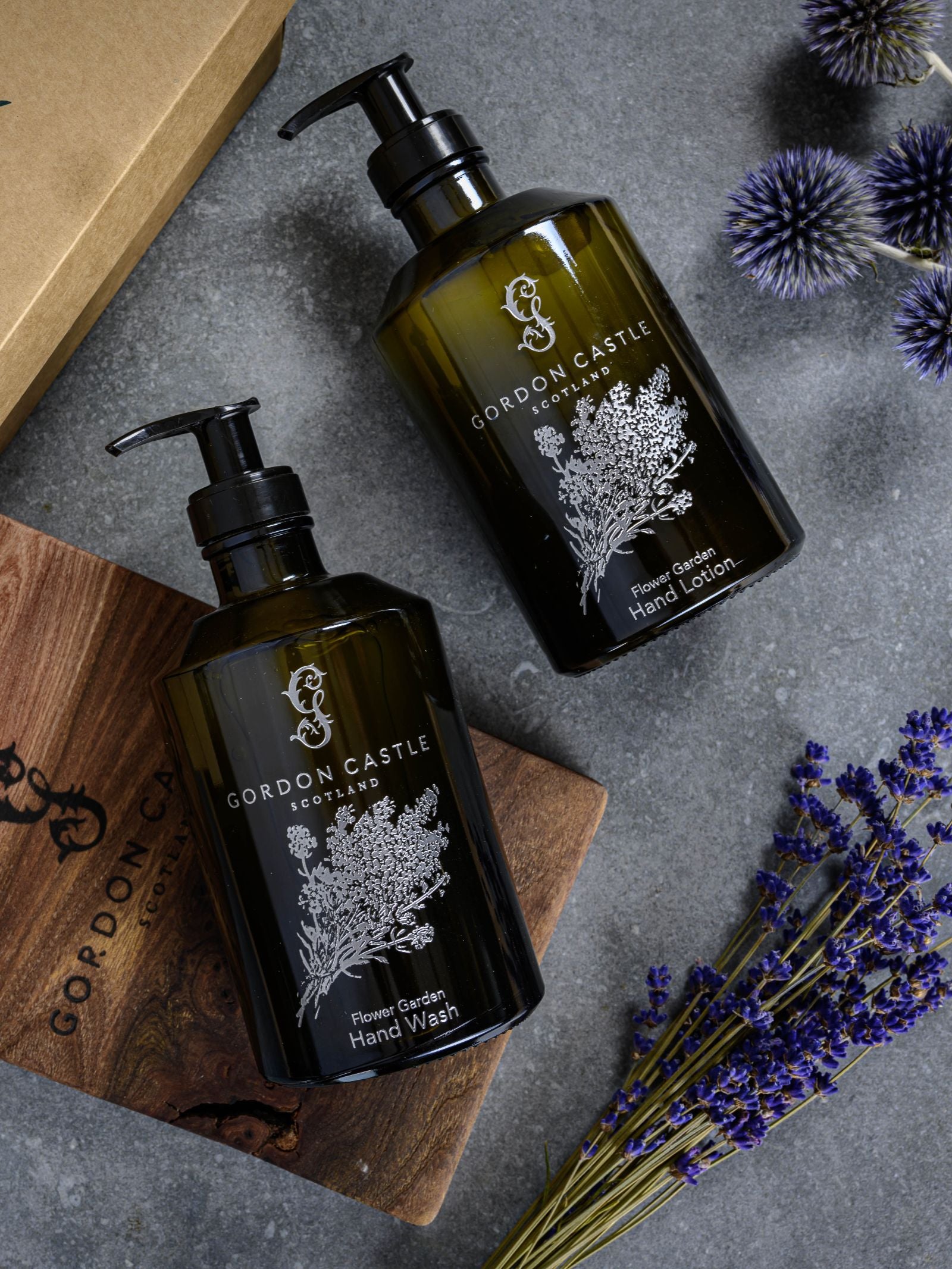 Two dark green refillable glass bottles of Gordon Castle's The Flower Garden Hand Wash sit on a gray surface, surrounded by a wooden board, purple thistles, and dried lavender.