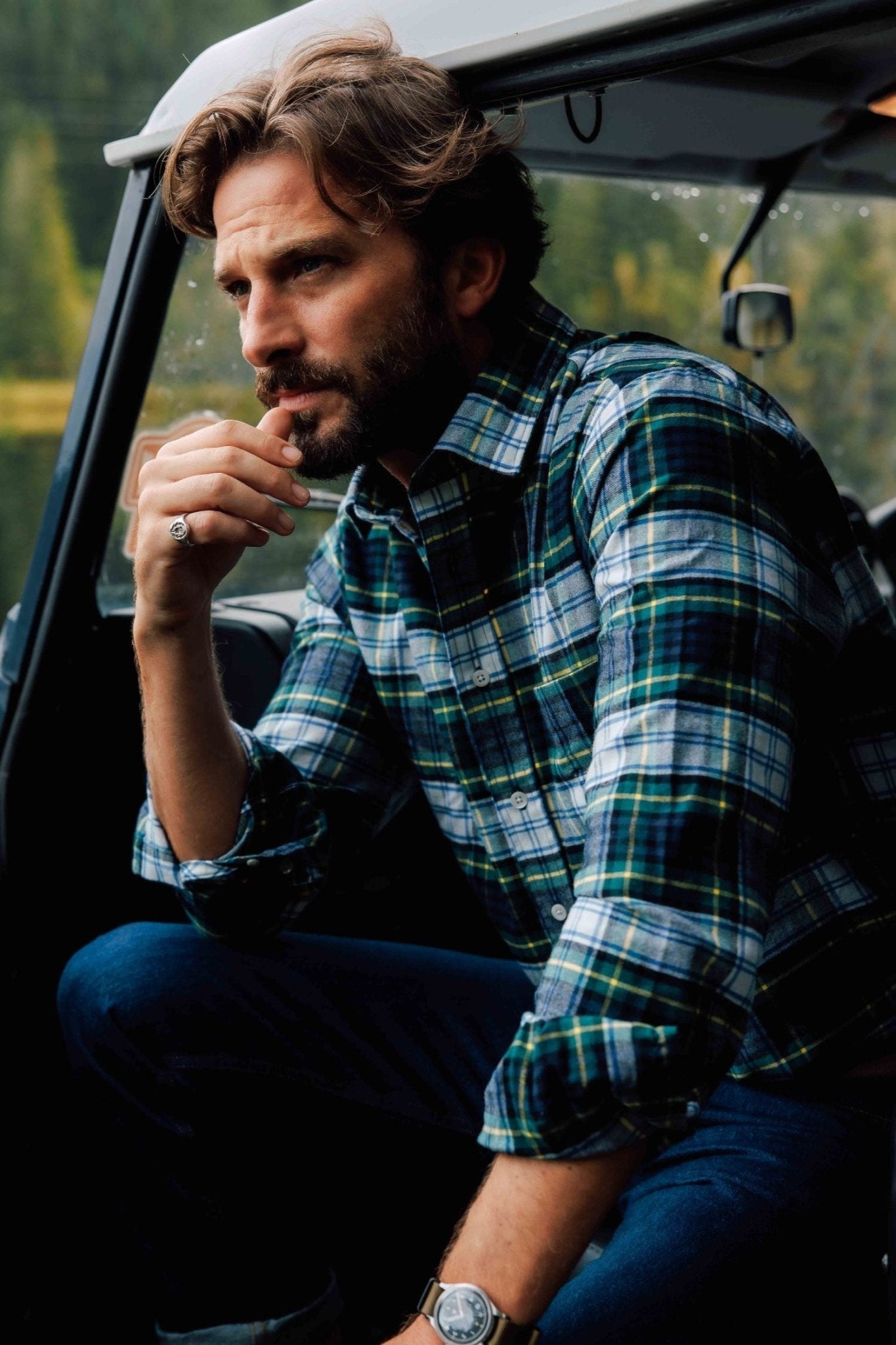 A bearded man with wavy hair sits in a vehicle, wearing Campbell's of Beauly Tartan Shirt in blue and green. He looks thoughtfully into the distance with one hand near his mouth, while forest scenery appears outside the window.