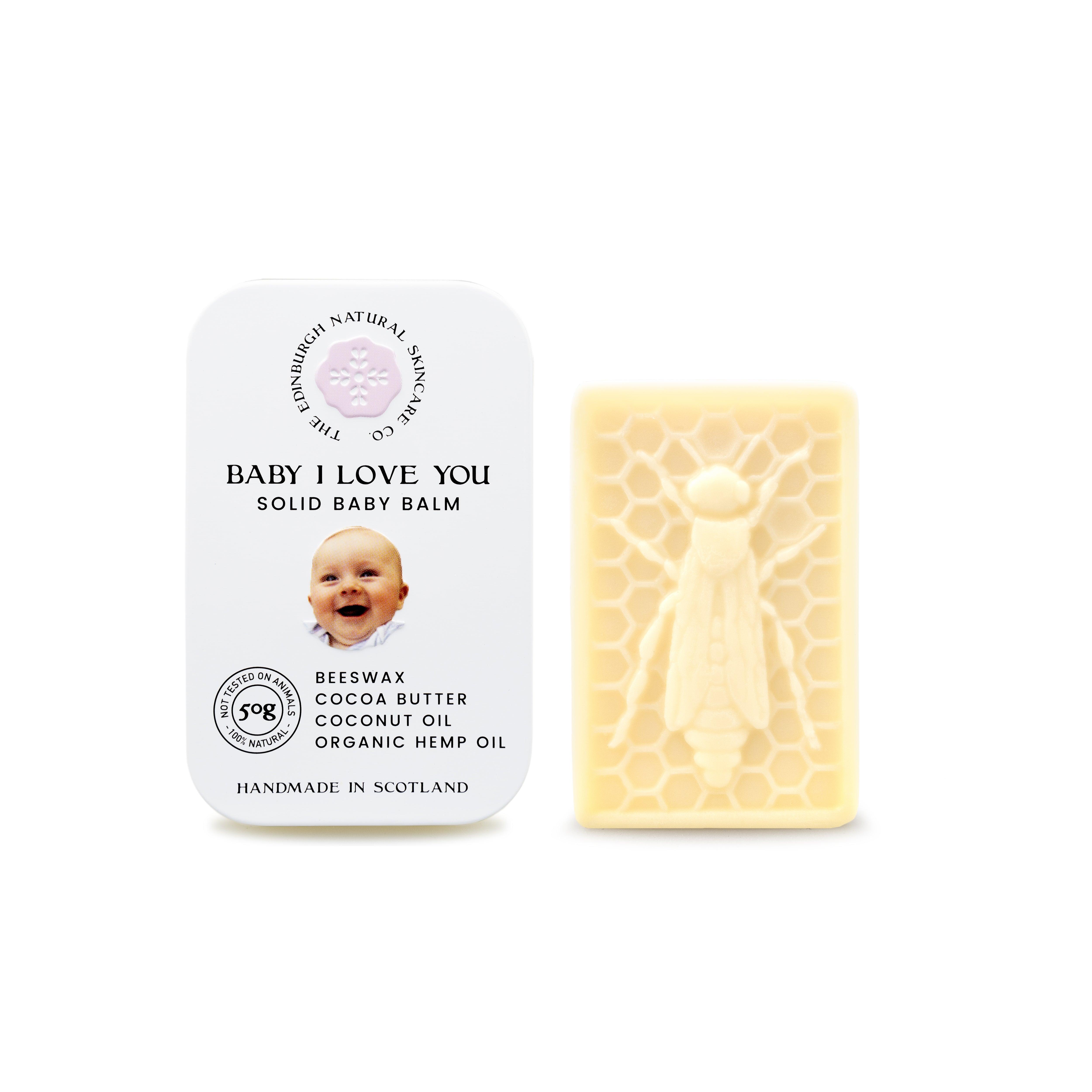 A white tin of Baby I Love You Solid Baby Balm by The Edinburgh Natural Skincare Company, featuring a smiling baby and skin-soothing oils, sits beside a yellow bee-shaped natural balm on a honeycomb pattern.