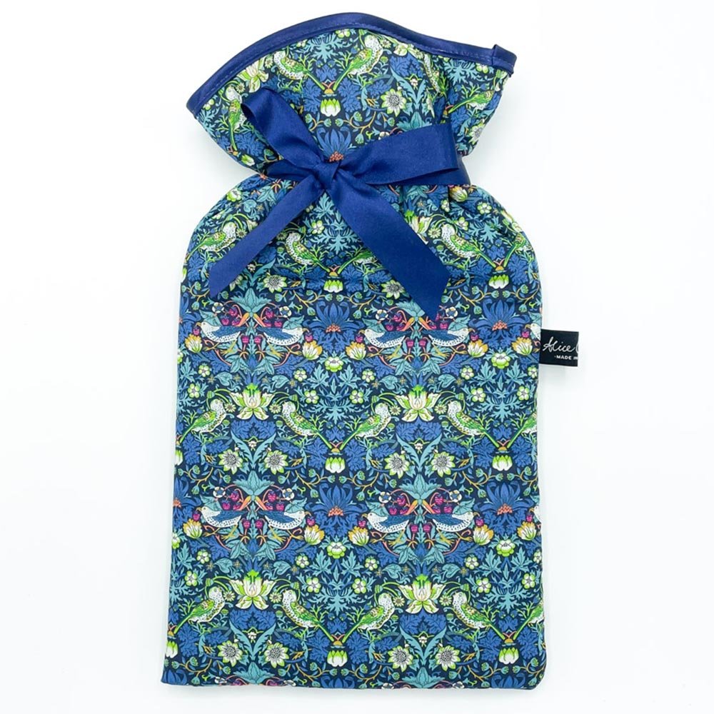 The Liberty Print Hot Water Bottle by Alice Caroline features blue Liberty cotton with a vibrant floral and bird design, finished with a dark blue ribbon bow. A black label with white text is attached to the side.