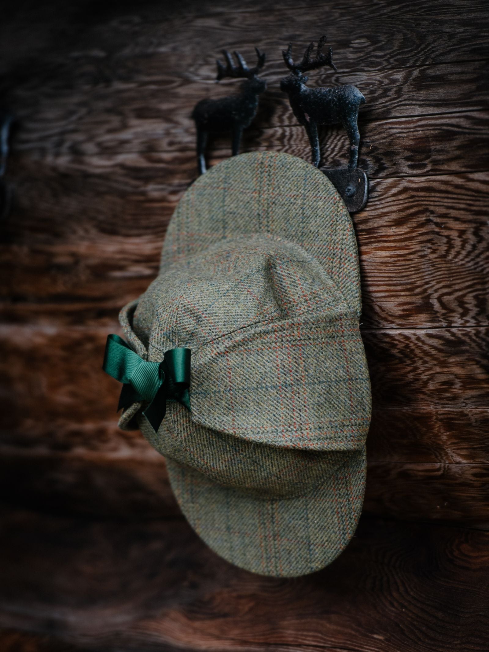 A Campbells of Beauly Sherlock Holmes Hat in green tweed, with earflaps and a green bow, hangs on a rustic wooden wall hook shaped like two deer.