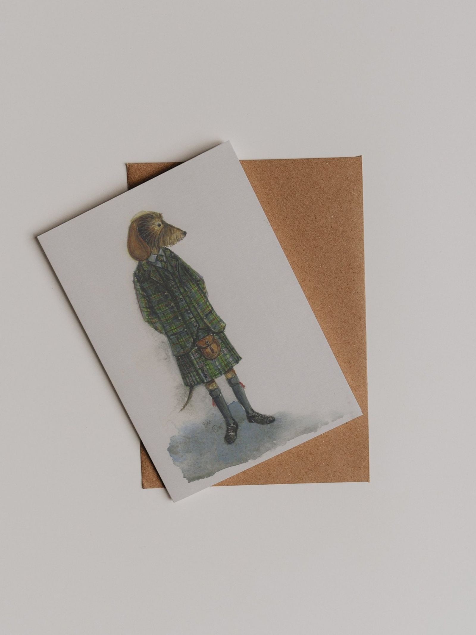 The Kilted Dachshund Greeting Card by Campbell’s of Beauly features an illustration of a dog in a green plaid coat standing upright, set atop a plain brown envelope against a white background. This charming thank you card adds whimsy and warmth to your thoughtful gesture.