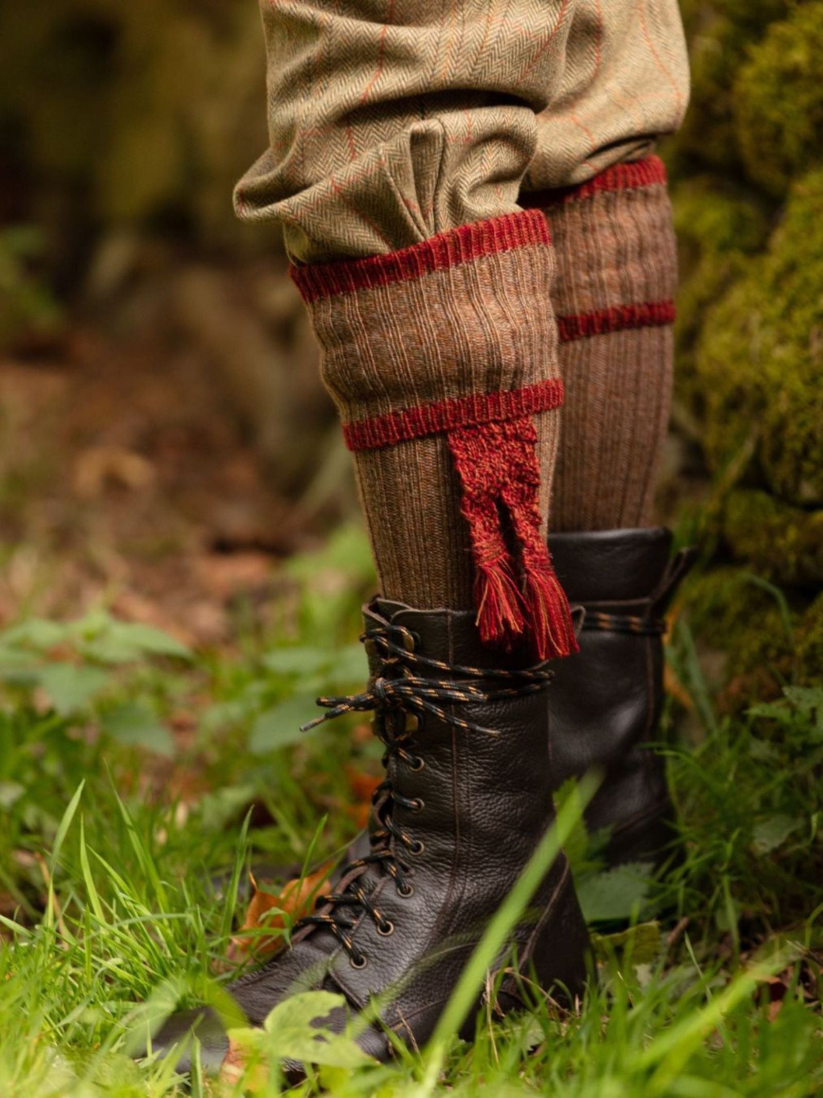 A close-up of a person’s lower legs wearing Campbell's of Beauly Kyle Socks with red cuffs and tassels, tan tweed trousers, and black lace-up boots, standing on green grass outdoors in the scenic Scottish Borders.