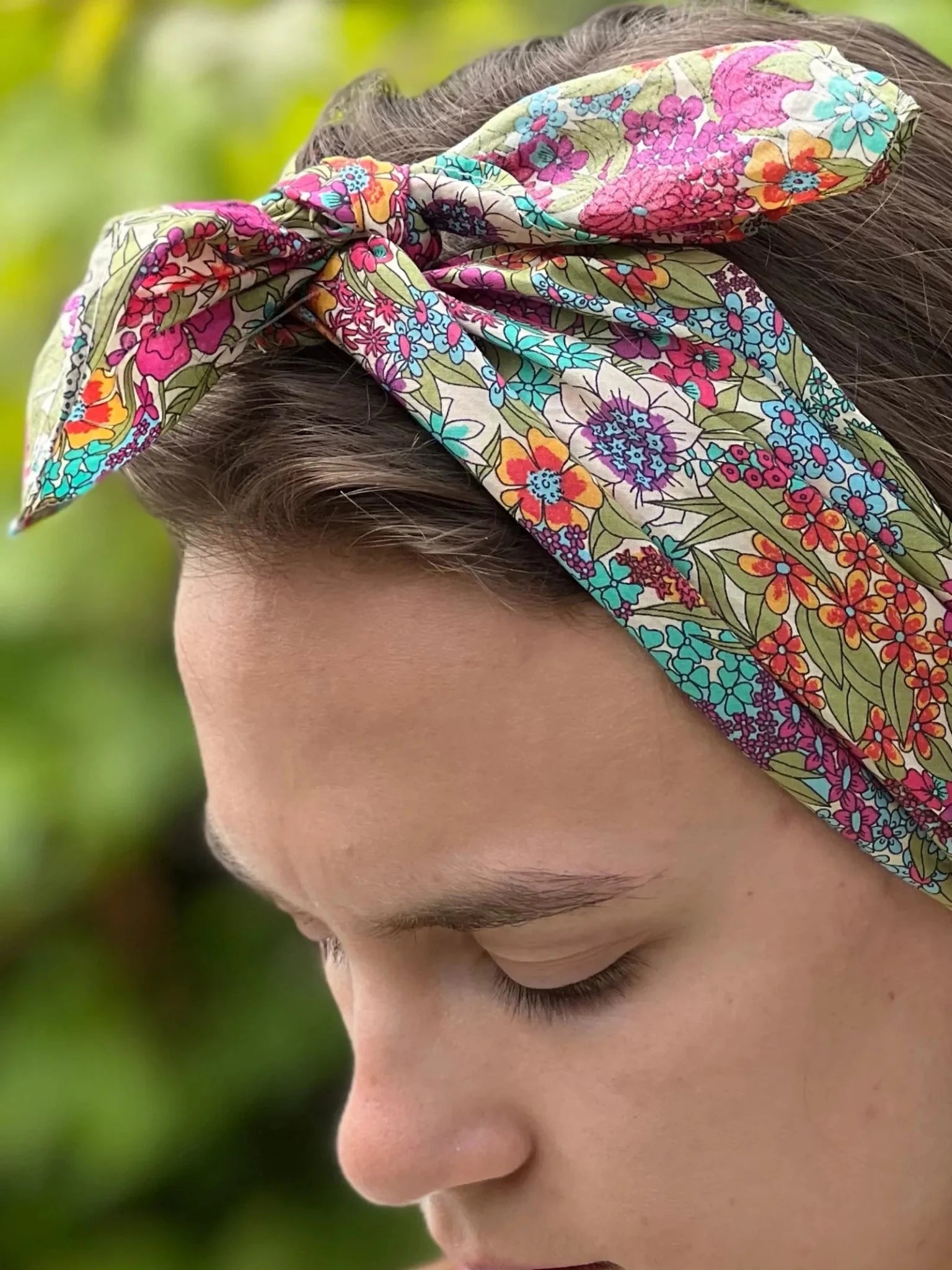 A close-up shows a person in profile wearing the Square Headscarf by Campbell's of Beauly, tied in a bow. The colorful floral pattern stands out against a blurred green foliage background.