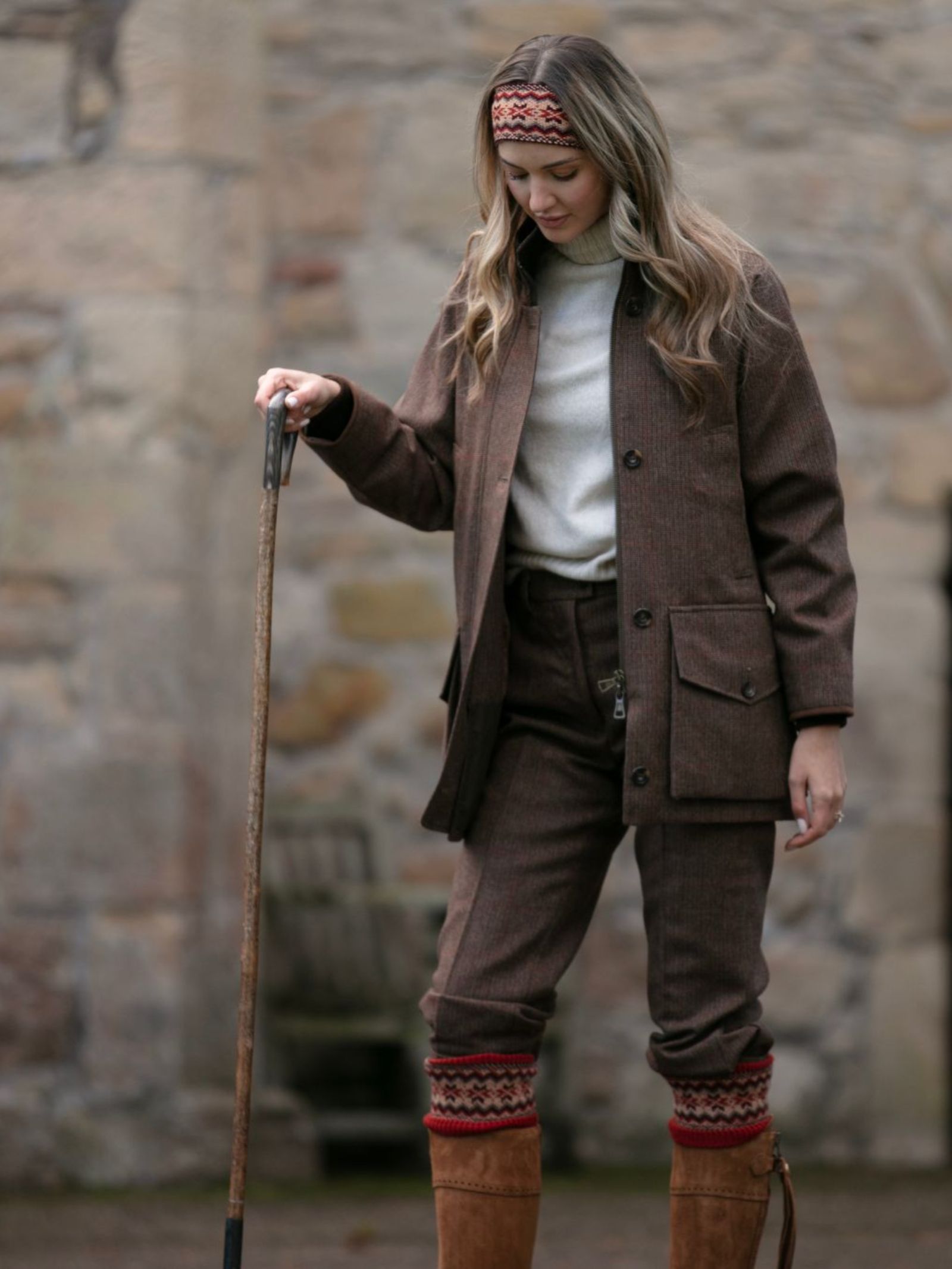 A woman outdoors in the Scottish Borders stands by a stone wall, wearing a Campbell's of Beauly Fairisle Headband, brown tweed jacket and pants, white turtleneck, tall brown boots, and patterned leg warmers while holding a walking stick.