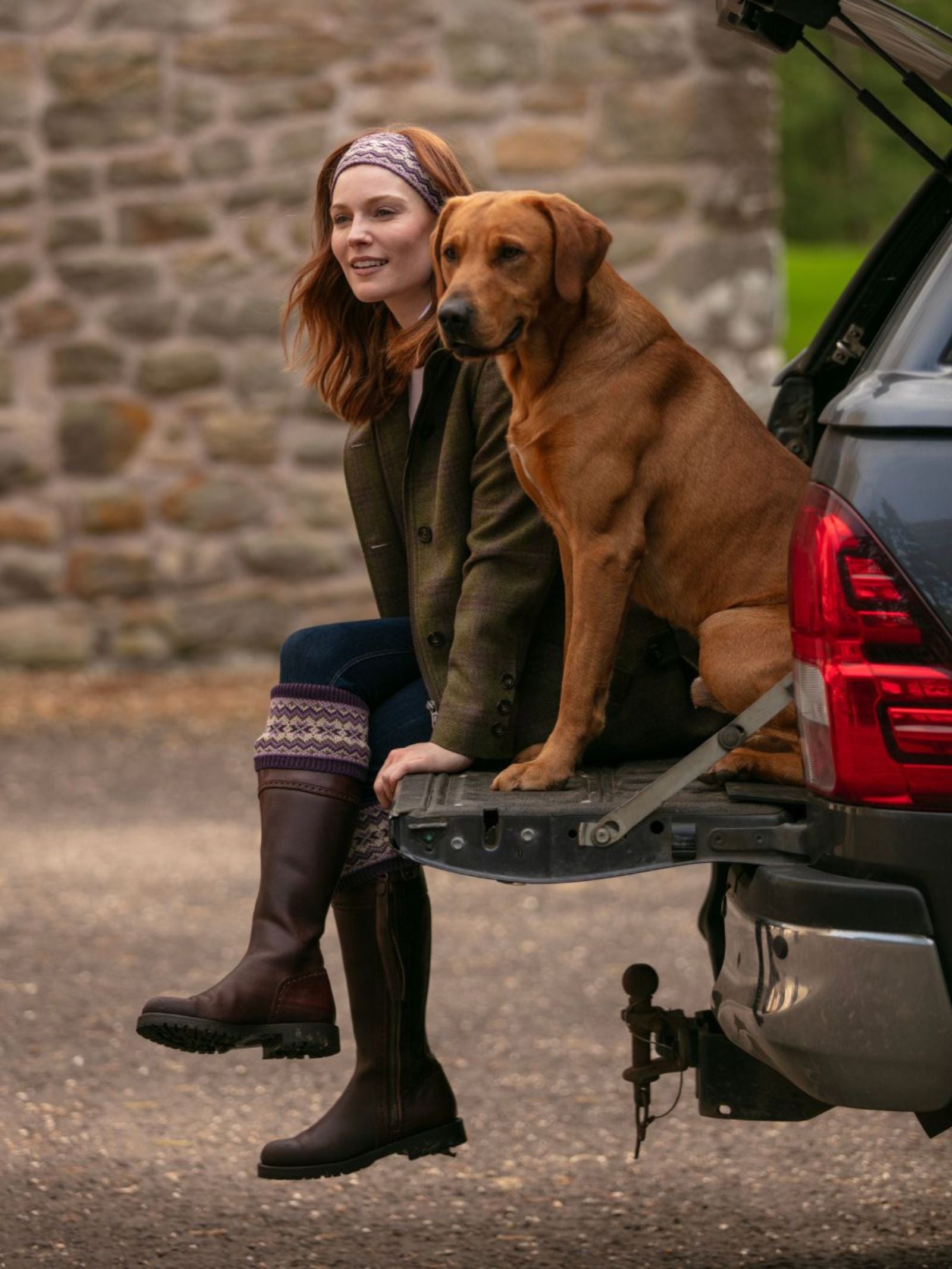 A woman wears a Campbell's of Beauly Merino Fairisle Shooting Socks patterned headband and sits on an open vehicle tailgate beside a large brown dog, both outdoors near a stone wall, looking ahead with relaxed expressions.