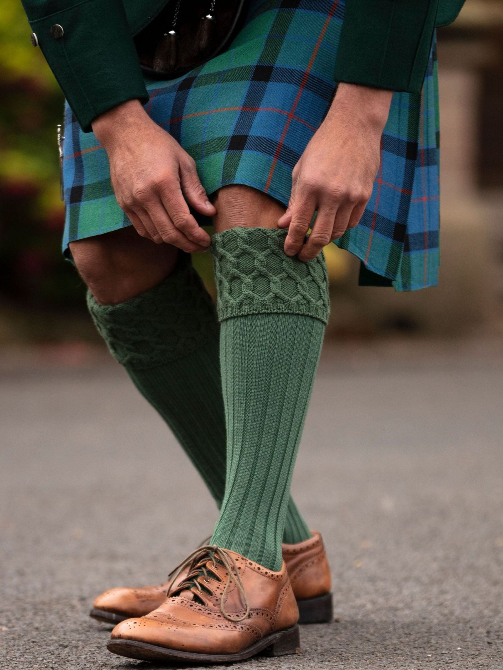 A person in Highland dress—wearing a blue and green tartan kilt, Campbell's of Beauly Kilt Socks, and brown leather brogues—stands outdoors adjusting a sock. Only their legs and hands are visible.