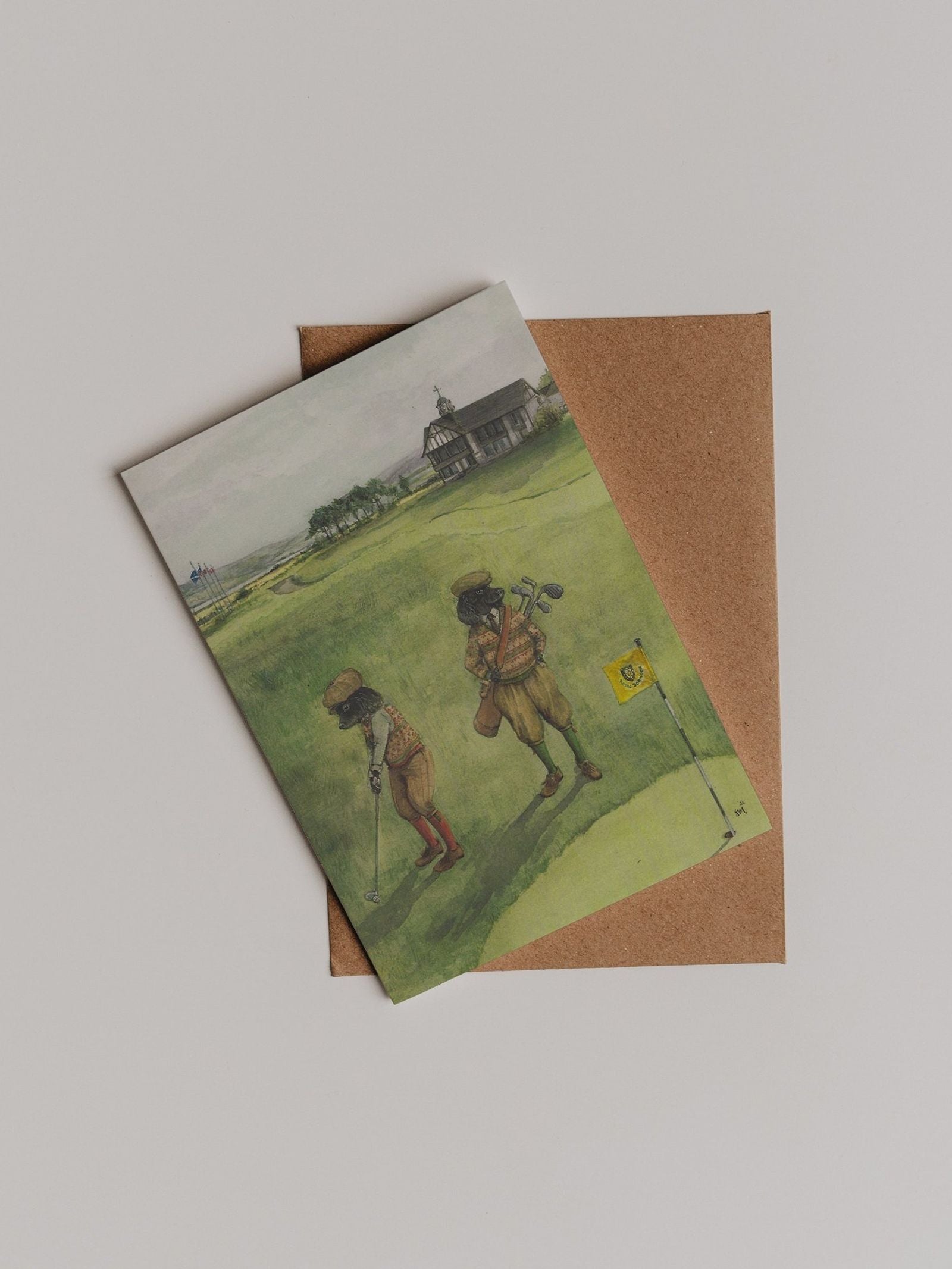 A Links Dornoch Greeting Card from Campbell’s of Beauly, featuring an illustration of two children golfing on green hills, is partially tucked into a brown envelope and displayed on a plain background.