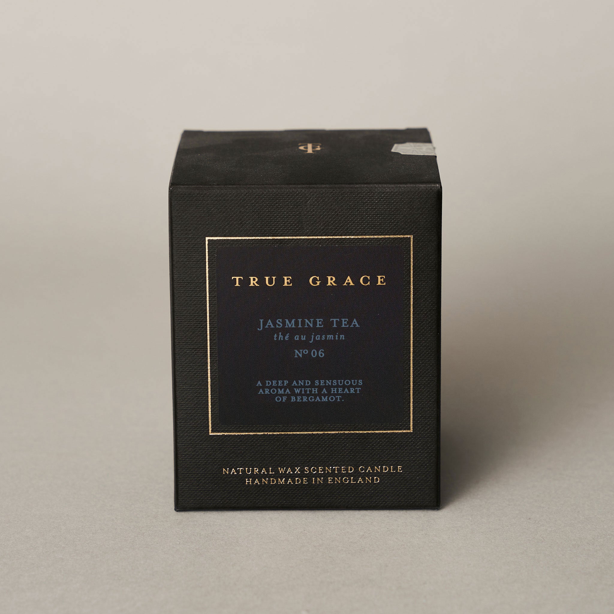A black box with gold and white text reads True Grace Jasmine Tea Candle, No. 06. This handmade candle from England blends rich jasmine and bergamot notes for a deep, sensuous scent. Made with natural wax.