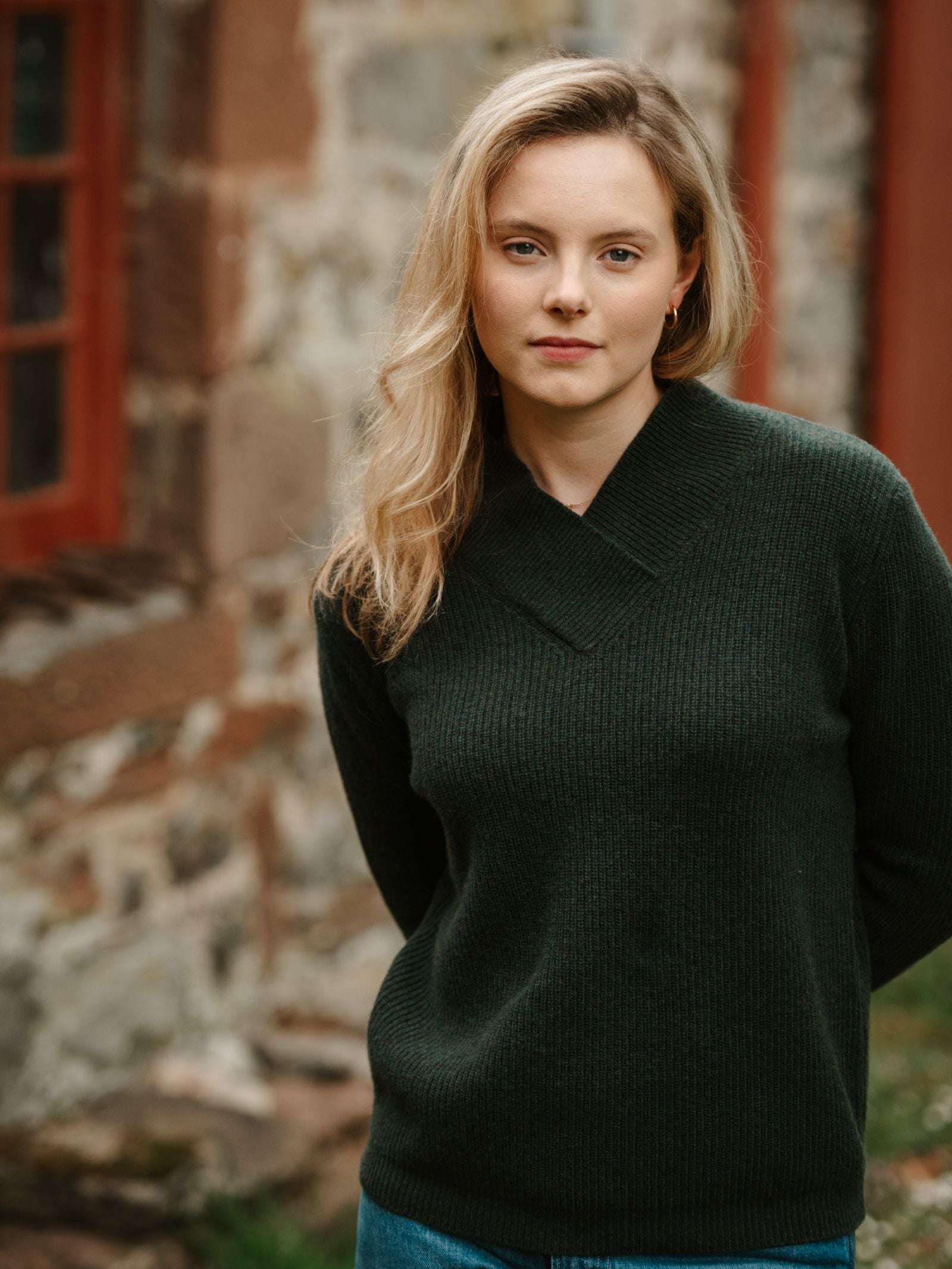 A woman with blonde hair wearing the Campbell’s of Beauly Rib V Neck Jumper in Dark Olive stands outside in front of a stone building with red window frames, looking at the camera with a neutral expression.