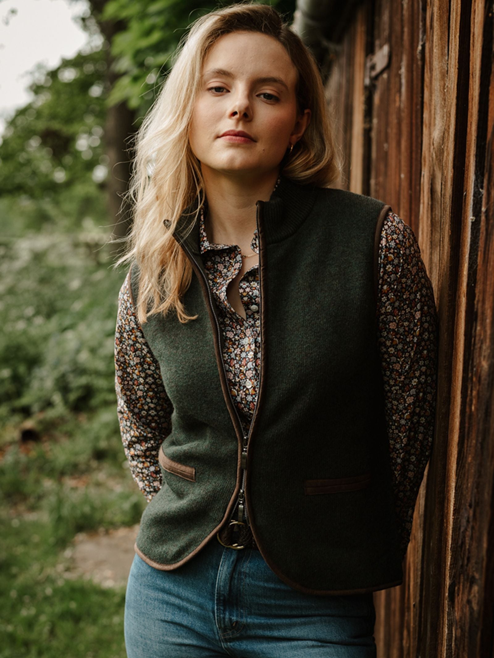 A woman with blonde hair wears a floral shirt, jeans, and the Merino Cashmere Gilet by Campbell’s of Beauly, standing outdoors by a wooden wall with greenery in the background, looking at the camera and effortlessly capturing timeless countryside style.