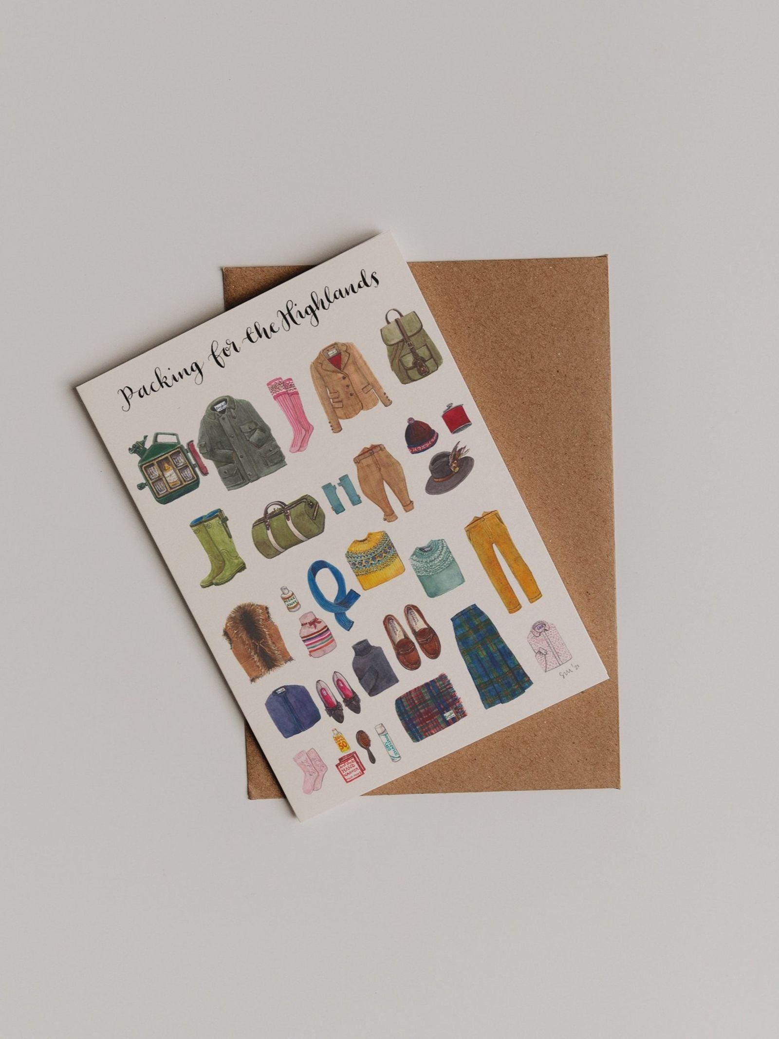 The Packing for the Highlands Greeting Card from Campbell’s of Beauly features illustrated clothing and travel essentials—such as shoes, boots, sweaters, jackets, pants, hats, backpacks, and accessories—in the brand’s classic style, displayed atop a brown envelope on a white background.