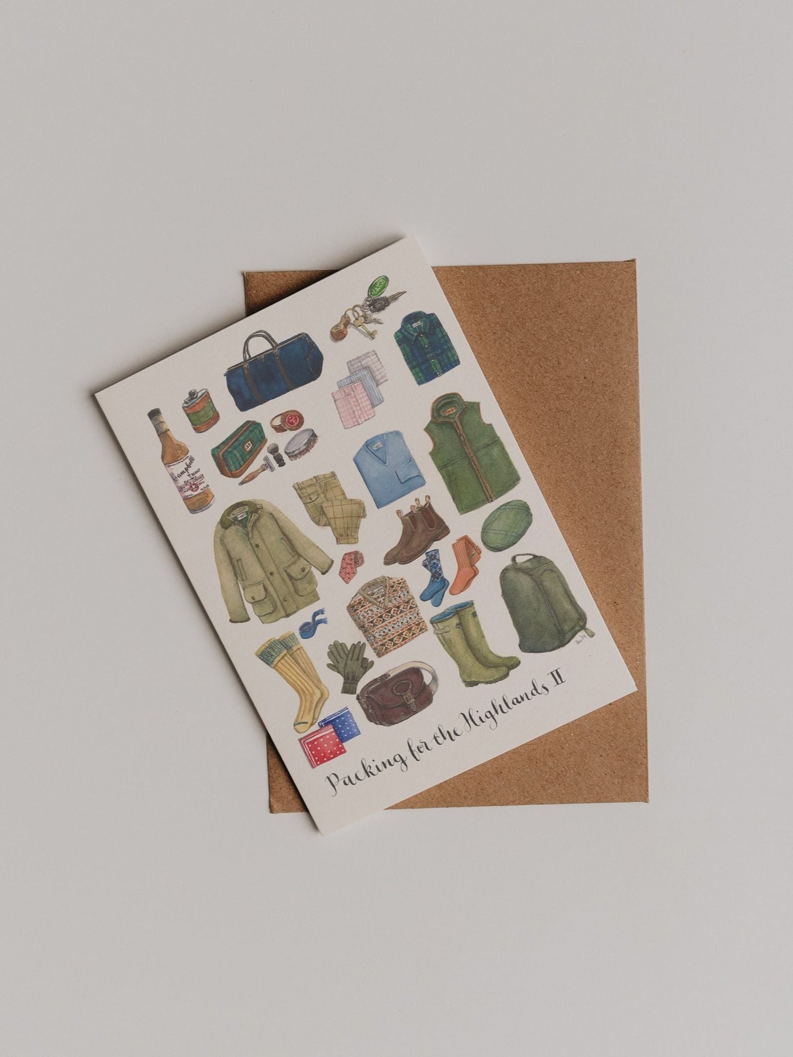 The “Packing for the Highlands 2 Greeting Card” by Campbell’s of Beauly features delightful travel and outdoor illustrations—including jackets, boots, bags, hats, and gifts—beautifully displayed on a thank-you card. The card rests on a brown envelope set against a plain light background.