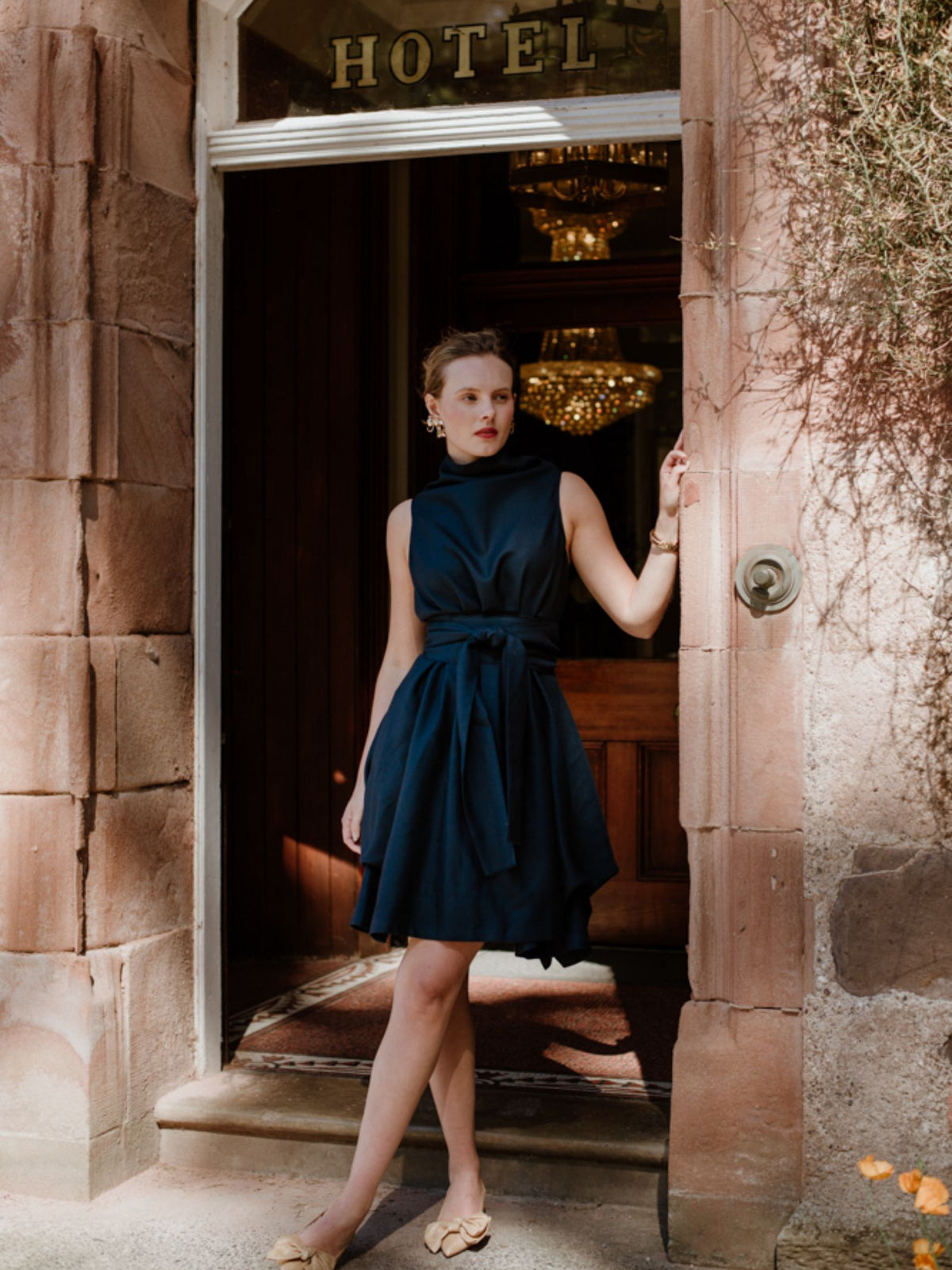 A woman in a sleeveless navy Campbell's of Beauly Reversible Wool Wrap Dress stands in a stone doorway beneath a HOTEL sign, one hand on the frame, gazing outside with chandeliers visible behind her.