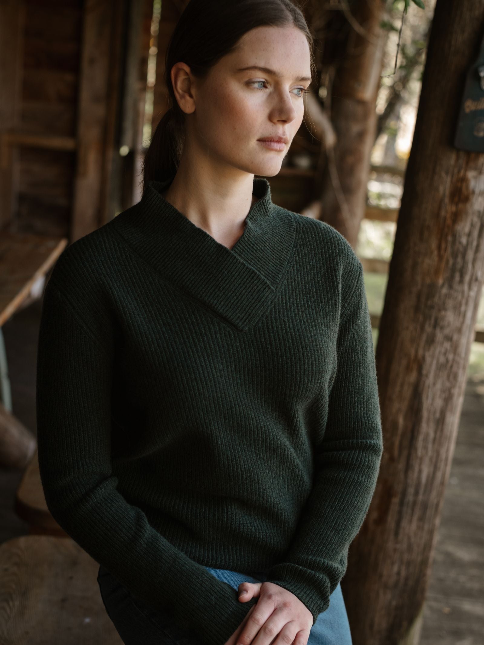 A woman with light skin and brown hair, wearing the Rib V Neck Jumper by Campbell’s of Beauly in dark green merino wool, sits indoors by wooden beams, looking thoughtfully to the side as natural light illuminates her face.