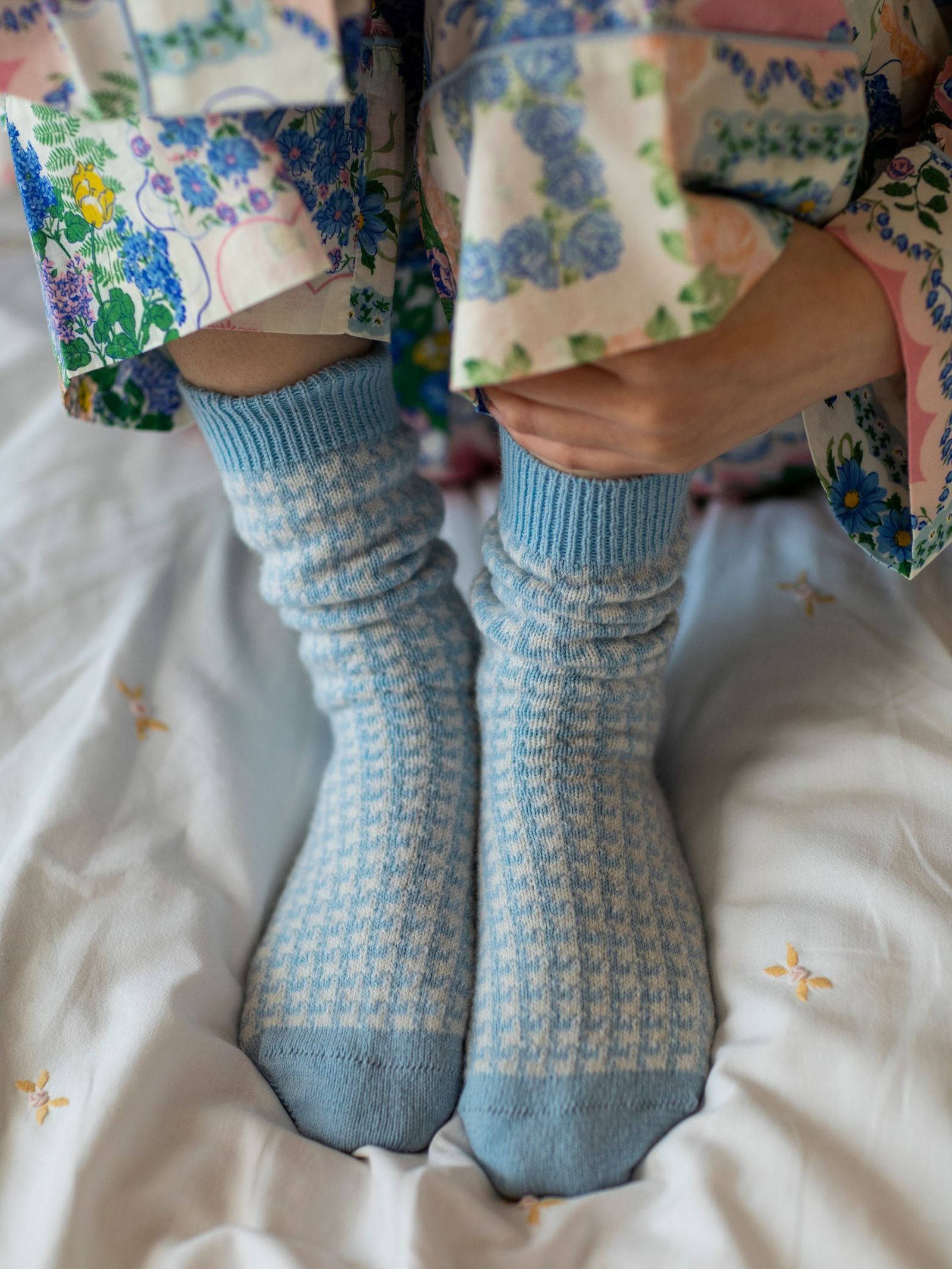 A person wearing Campbell’s of Beauly Cashmere Houndstooth Socks in blue sits on a bed with a white floral duvet, dressed in colorful, floral-patterned pajamas and holding their knees.