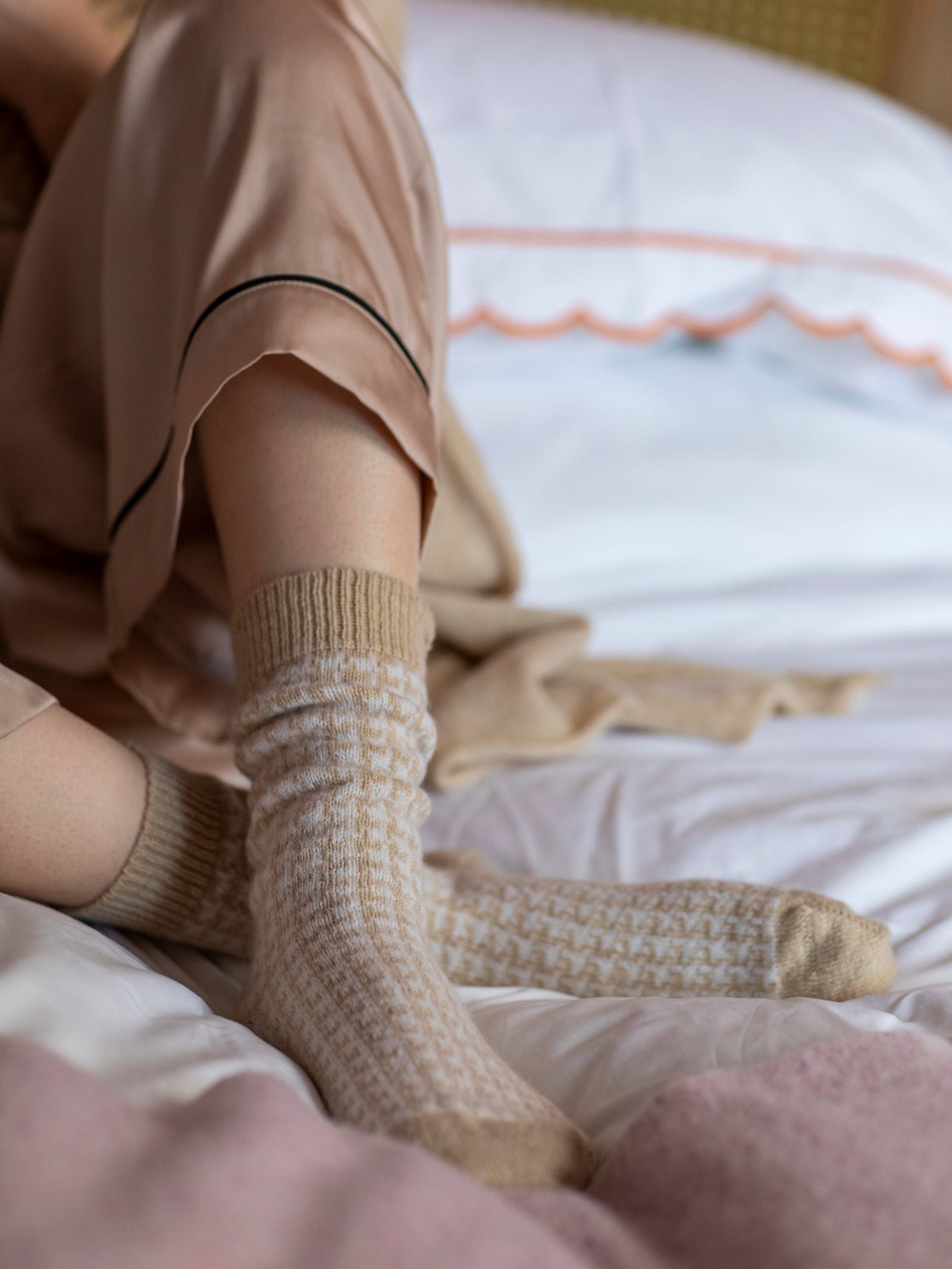 A person wearing Campbell’s of Beauly Cashmere Houndstooth Socks in Natural sits on a bed with white and pink bedding, dressed in beige pajamas. Only the lower half of their body is visible.