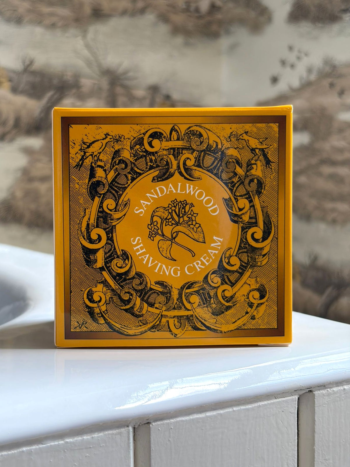 A yellow and brown box of Geo. F. Trumper Sandalwood Shaving Cream sits on white tiles by a sink, nature-themed wallpaper behind it—this gentle, glycerine-based cream is ideal for sensitive skin.