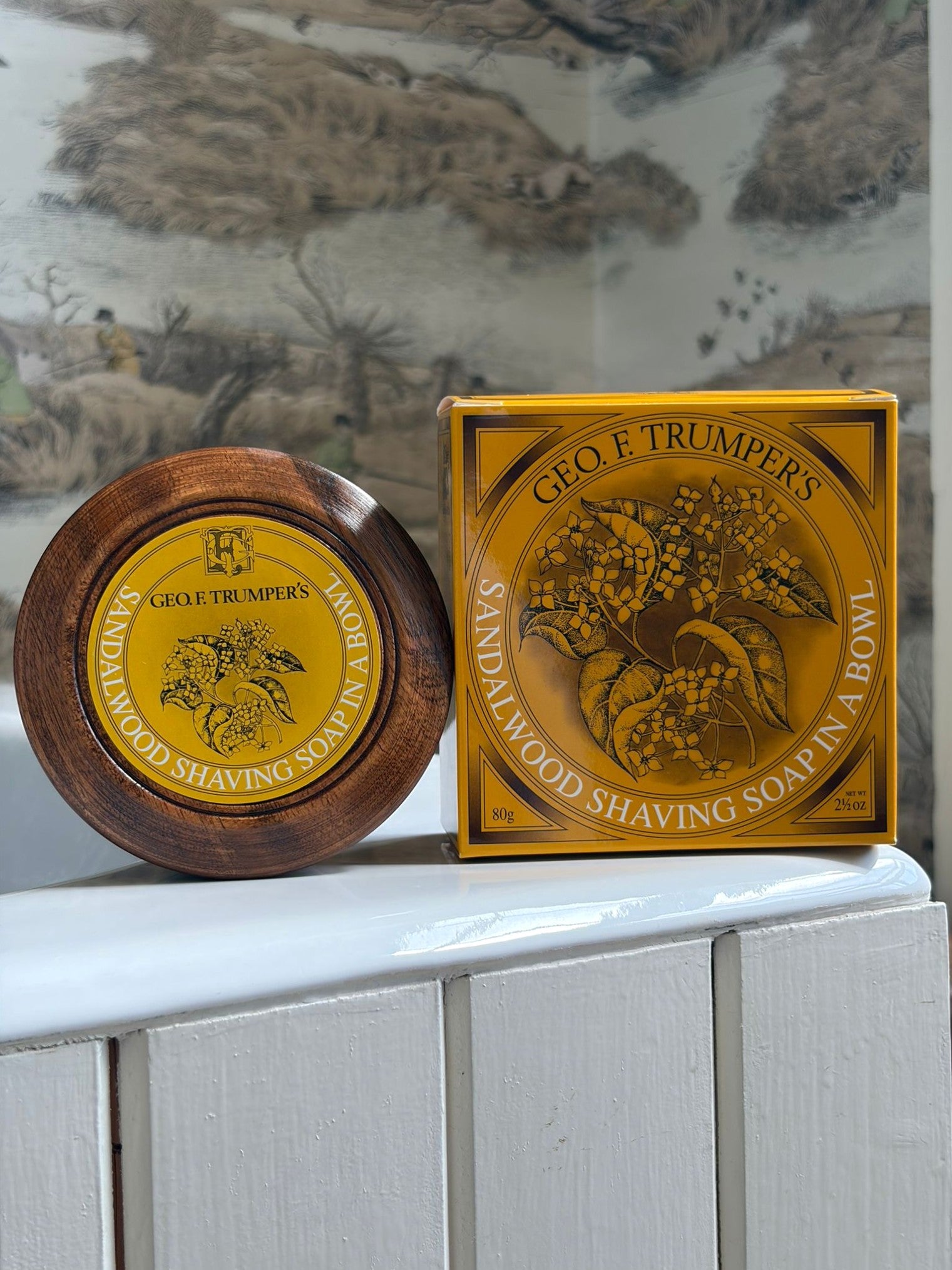 A wooden bowl and a yellow box labeled Geo. F. Trumper Sandalwood Shaving Soap & Bowl rest on a white wooden surface with a decorative backdrop—ideal for an elevated shaving experience.