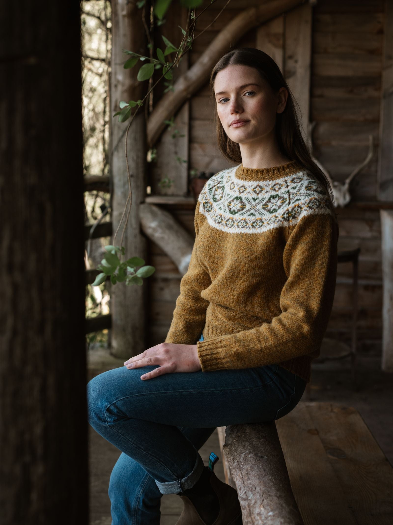 A woman with long brown hair, wearing the Campbell's of Beauly Shetland Fairisle Crew in mustard yellow and white with blue jeans, sits on a wooden beam in a rustic wooden cabin with antlers in the background.