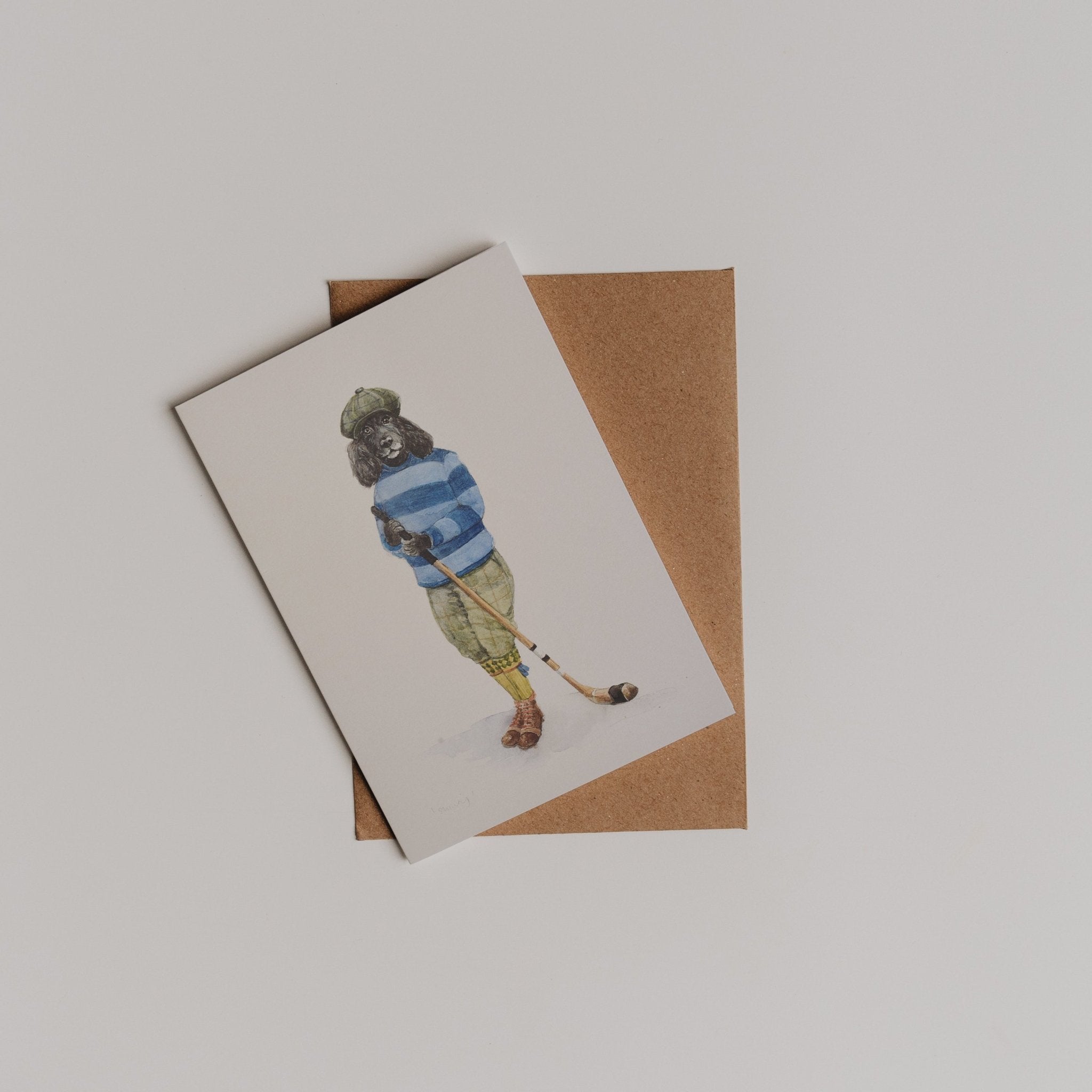 The Shinty Player Greeting Card by Campbell's of Beauly, featuring a dog in vintage golfing attire, is shown atop a brown envelope on a plain white background.