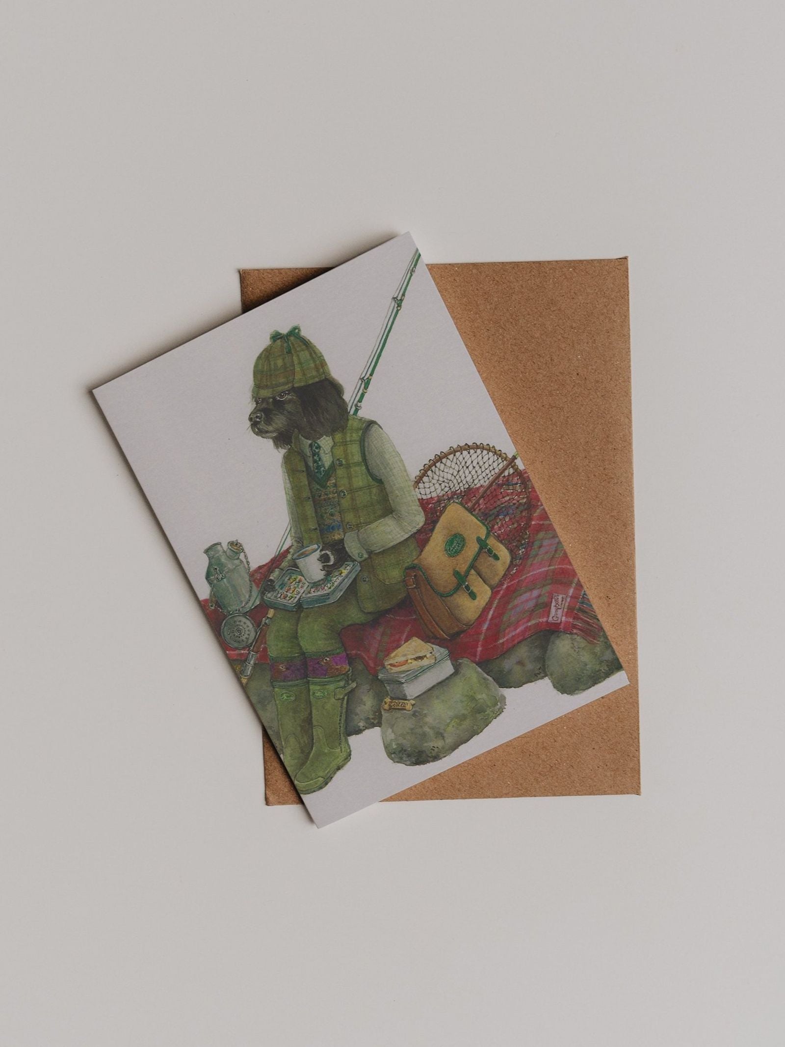 The Fisherman Greeting Card by Campbell’s of Beauly showcases a charming Sophie Murray illustration of a dog dressed in fishing gear, seated on rocks with a cup in hand, accompanied by fishing equipment and a bag—all presented with a classic brown envelope, making it an ideal thank you note.