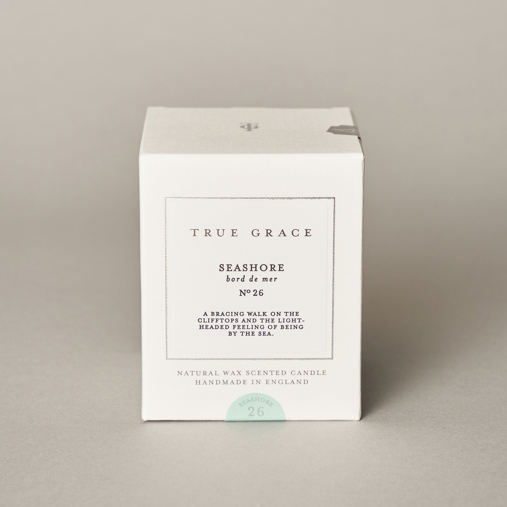 A white box labeled True Grace Seashore Candle stands on a neutral background. The label describes its marine scent as A bracing walk on the clifftops and the light-headed feeling of being by the sea. Handmade in England.
