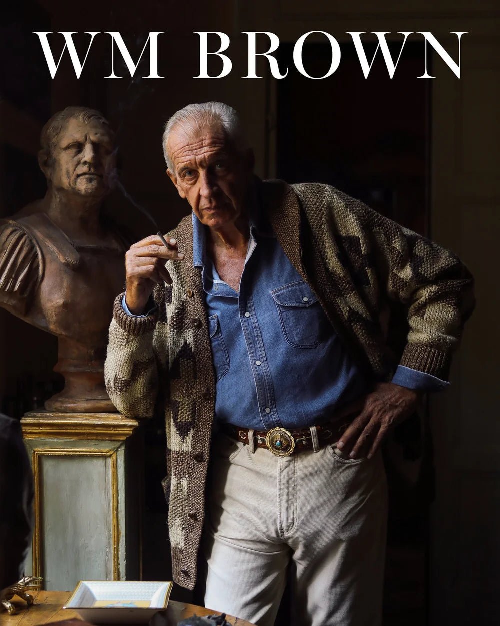 An older man in a denim shirt and patterned cardigan stands by a classical bust, holding a cigar. His vintage style echoes the essence of WM Brown, featured on the cover of William Brown Issue No 21 with bold white lettering.