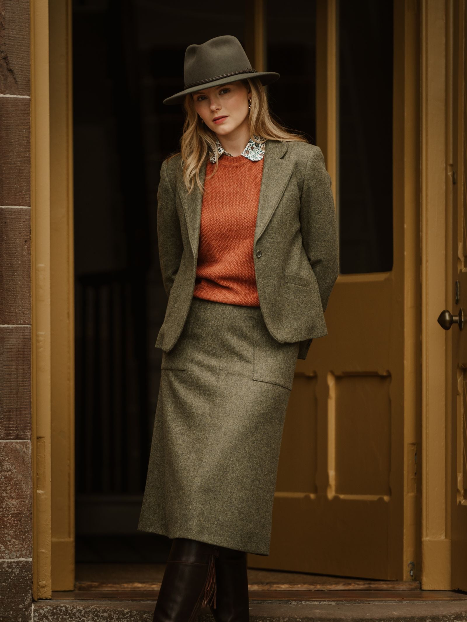 A woman in a Campbell's of Beauly House Jacket, styled with a green skirt, orange sweater, bold necklace, and hat, stands before a yellow wooden door. Her blonde hair is loose and she has a neutral expression.