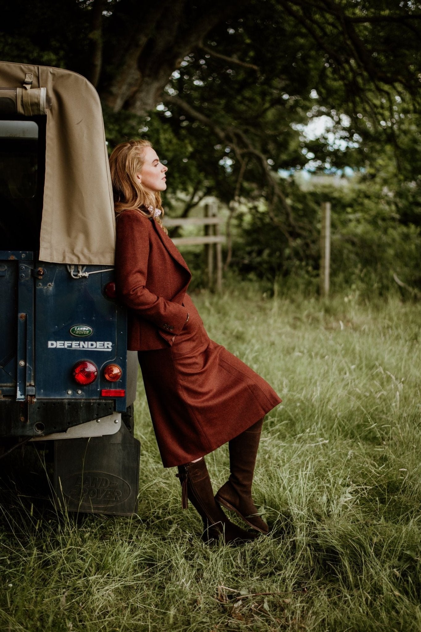 A woman in a brown A-Line Skirt by Campbell's of Beauly and boots leans against the back of a blue Land Rover Defender in a grassy field, with trees and a wooden fence in the background.