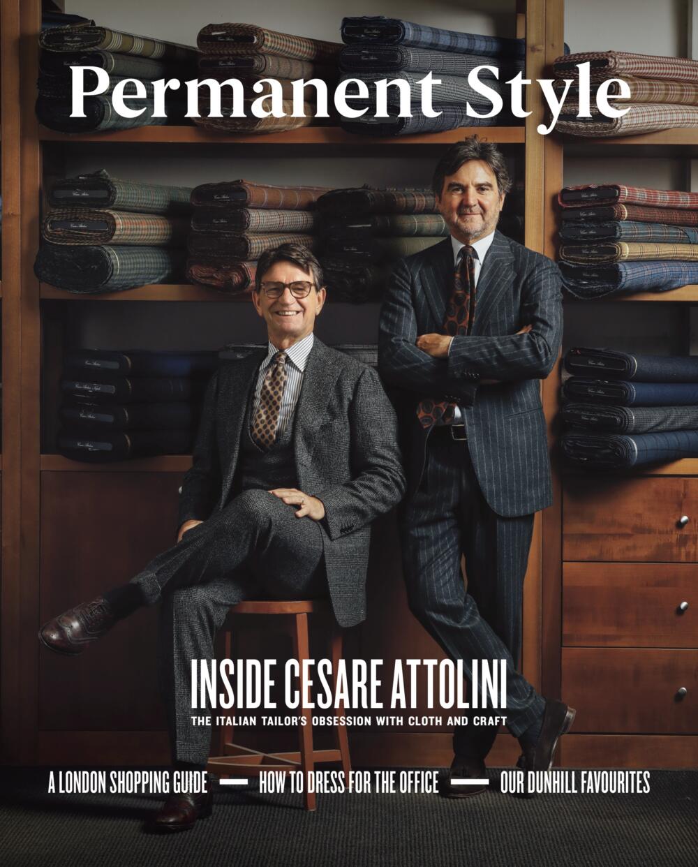 Two men in tailored suits pose before shelves of fabrics—one seated, one standing—in a formal scene that captures classic menswear and tailoring featured in Permanent Style Magazine, Issue 2 by Permanent Style.