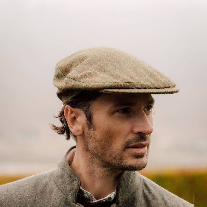 A man wearing the Brown Beauly Cap by Campbells of Beauly, styled in classic British fashion, pairs it with a gray jacket outdoors. He has short dark hair, a neat beard, and gazes slightly right against a blurred background.