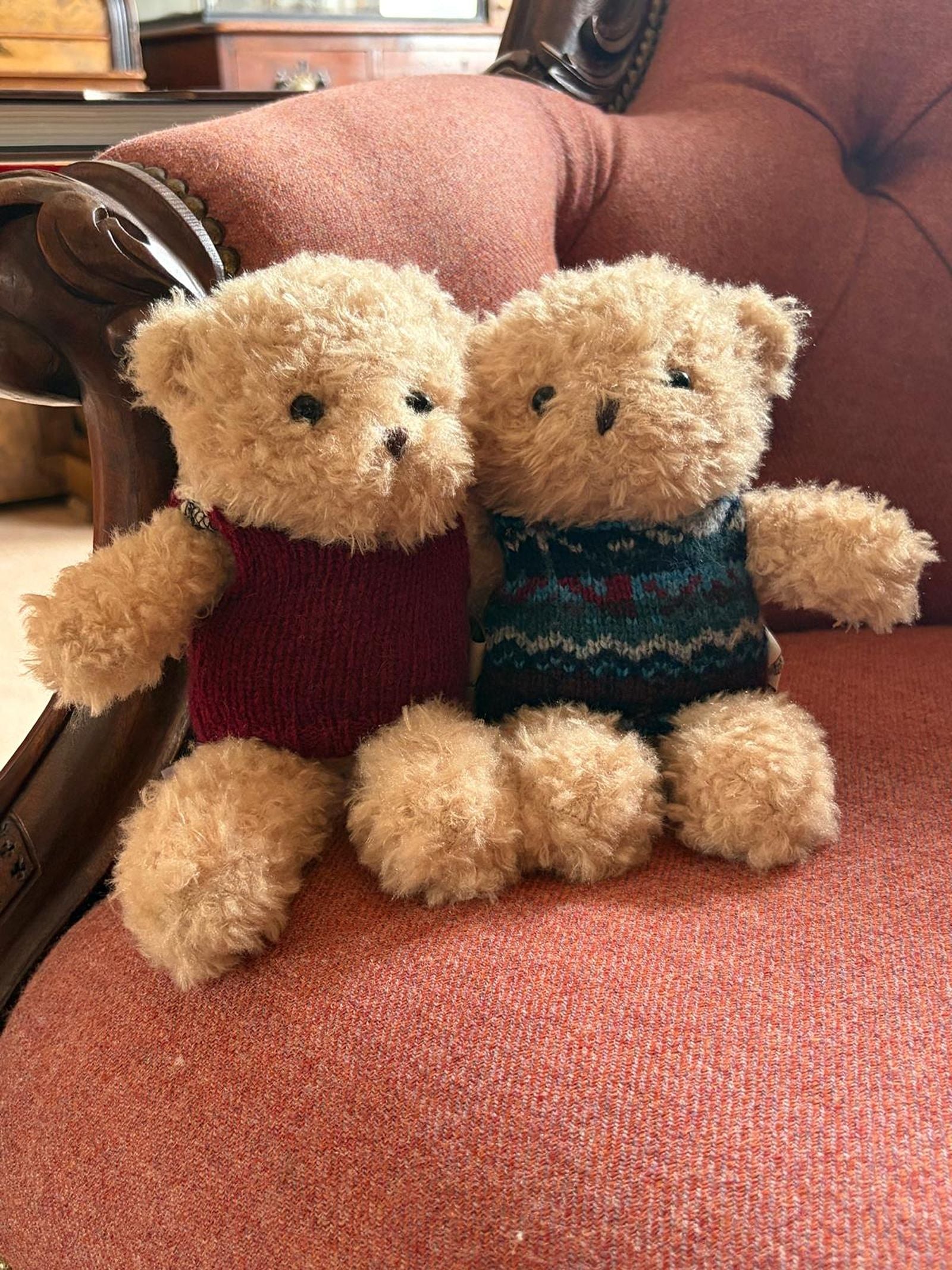 Two plush Bobby the Bear toys from Campbell’s of Beauly sit side by side on an orange upholstered chair. One wears a red Campbell’s of Beauly sweater, while the other sports a blue patterned Campbell’s knitwear jumper. With their button eyes and stitched noses, they appear irresistibly soft and cuddly, perfectly capturing the charm of these delightful bears by Campbell’s of Beauly.