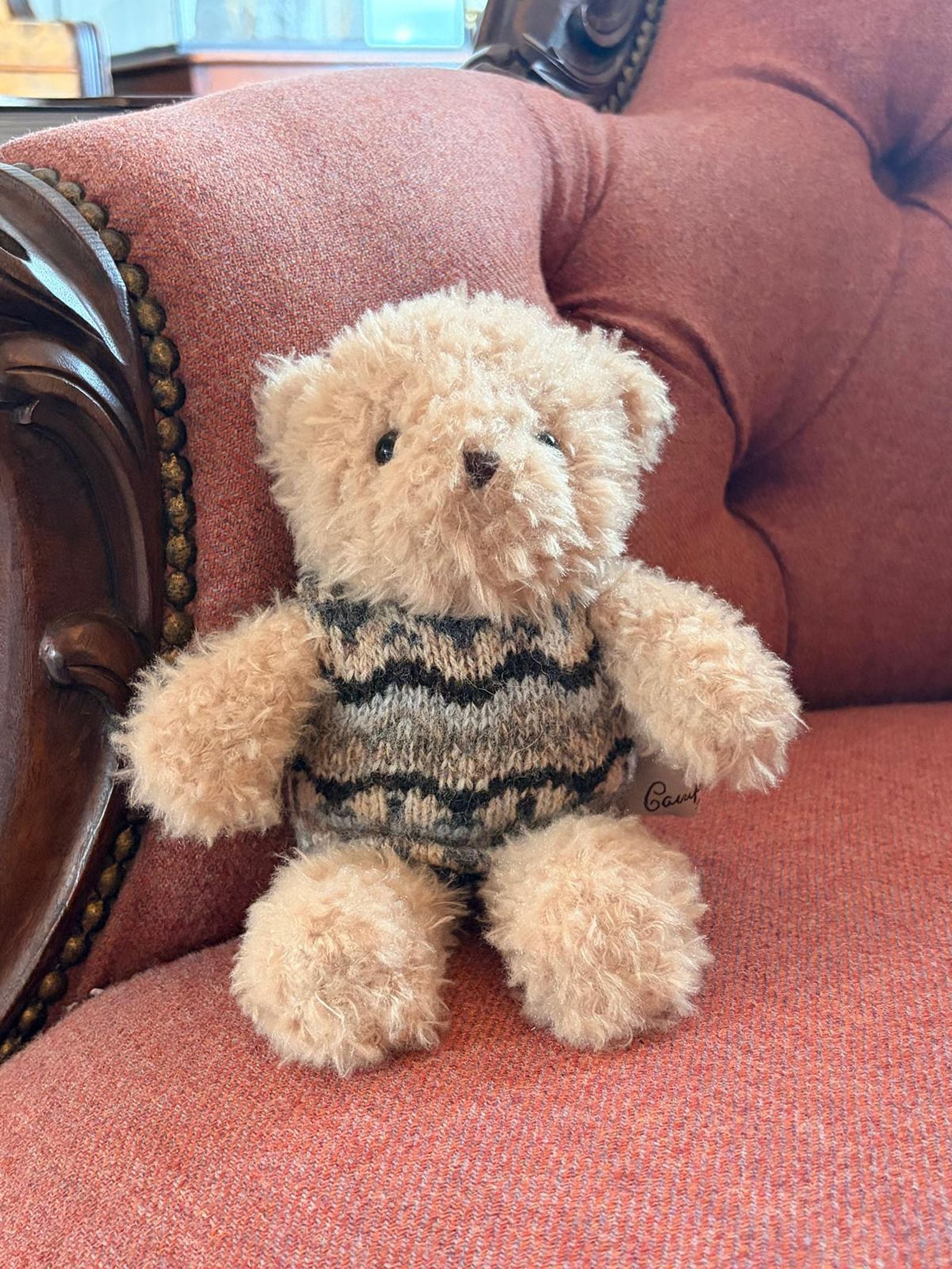 Bobby the Bear, a small and fluffy teddy bear from Campbell’s of Beauly, sits comfortably on an ornate red upholstered armchair, dressed in a stylish gray and black Campbell’s knitwear sweater.