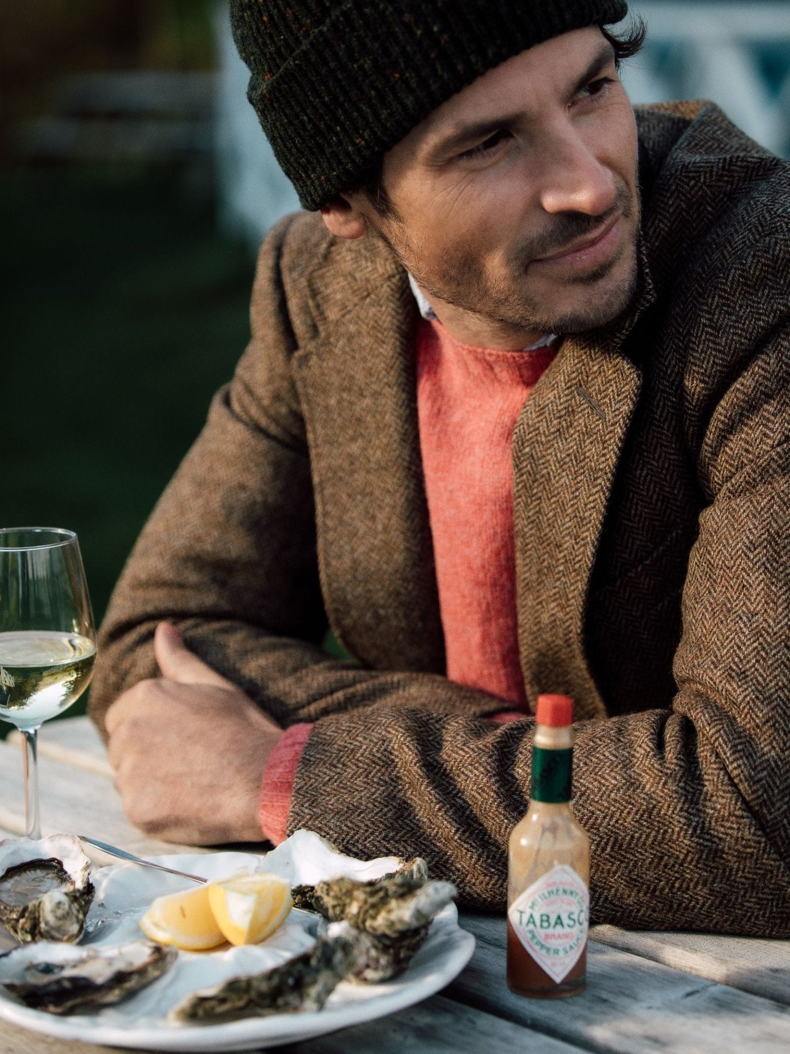 Wearing a Campbell's of Beauly Brown Herringbone House Jacket and a black knit hat, a man sits outdoors at a table with oysters, lemon wedges, Tabasco sauce, and white wine, looking to the side with a slight smile.
