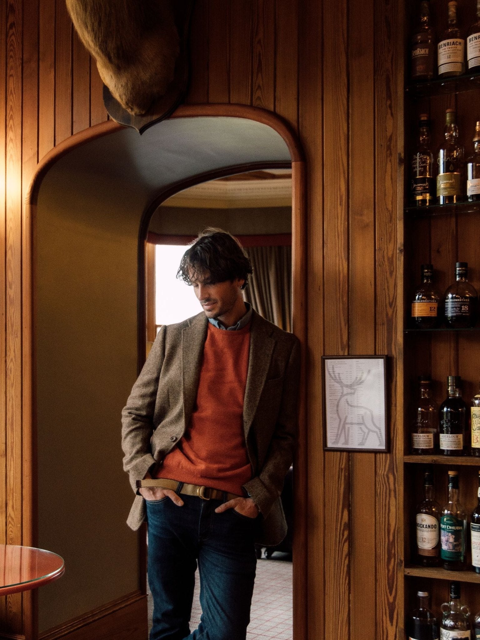 A man wears a Brown Herringbone House Jacket by Campbell's of Beauly and an orange sweater, standing by a wood-paneled wall near liquor shelves, hands in pockets, with a framed deer print and mounted antlers in view.