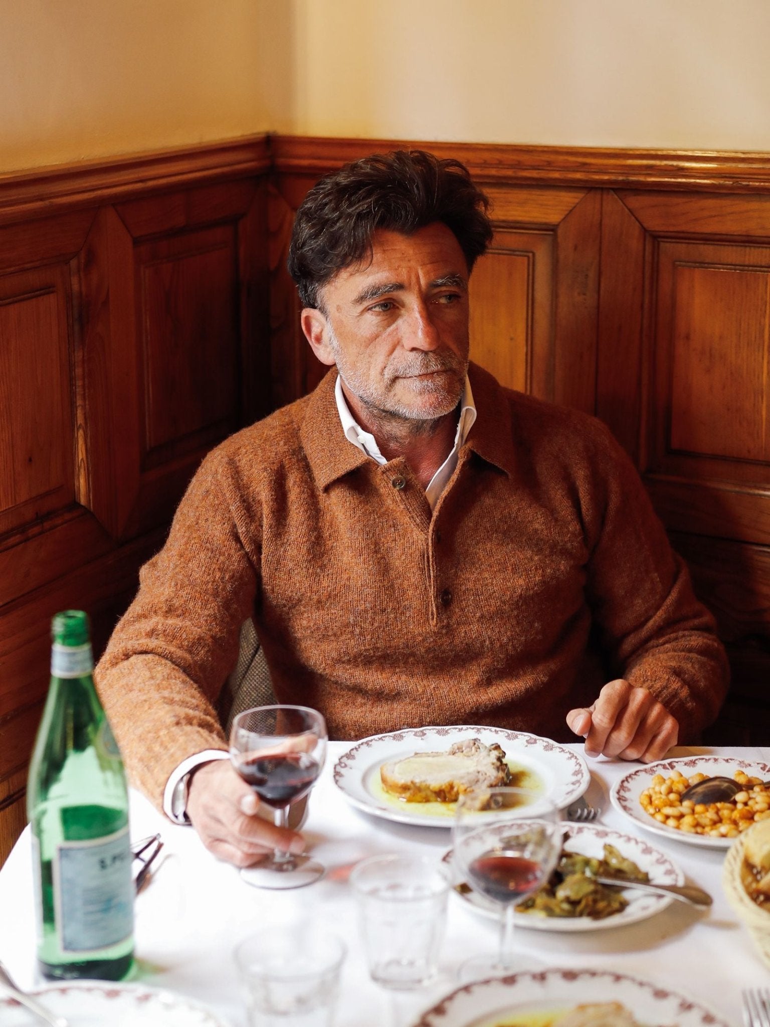 A man with dark hair wearing a Campbell’s of Beauly Brushed Shetland Rugby Jumper sits at a restaurant table with plates of food, holding a glass of red wine. A green bottle of water and wood-paneled walls are visible in the background.