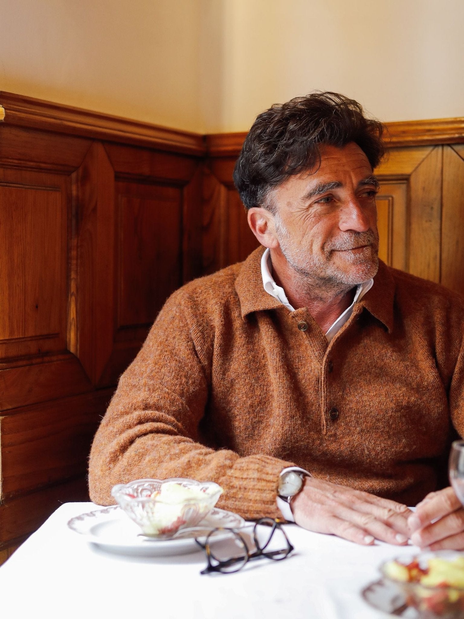 A man with dark hair and a beard, wearing the Brushed Shetland Rugby Jumper by Campbell’s of Beauly, sits at a table in a warm wooden booth, smiling and looking to the side. Glasses, a watch, and a dessert are visible on the table.